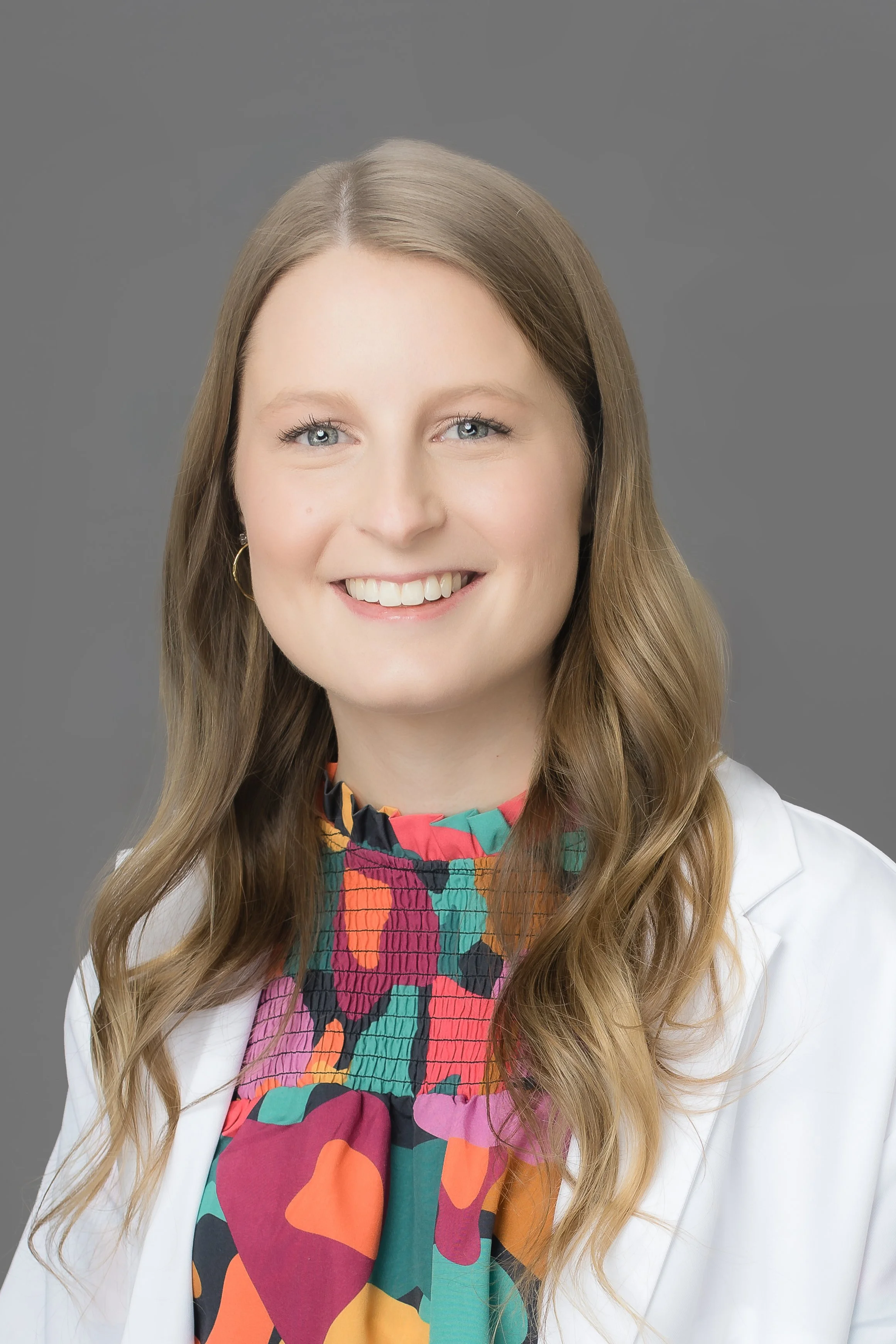 Portrait of a woman with long wavy blonde hair, blue eyes, smiling, wearing a colorful patterned blouse and a white blazer, against a gray background.