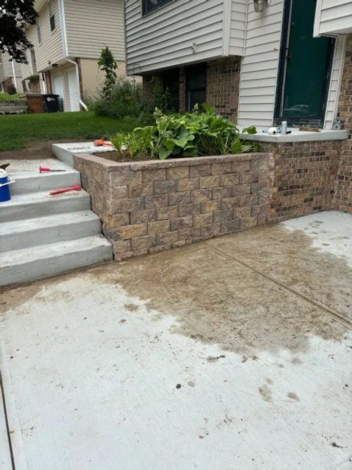 Newly built brick planter with green plants next to concrete stairs and sidewalk outside a house.