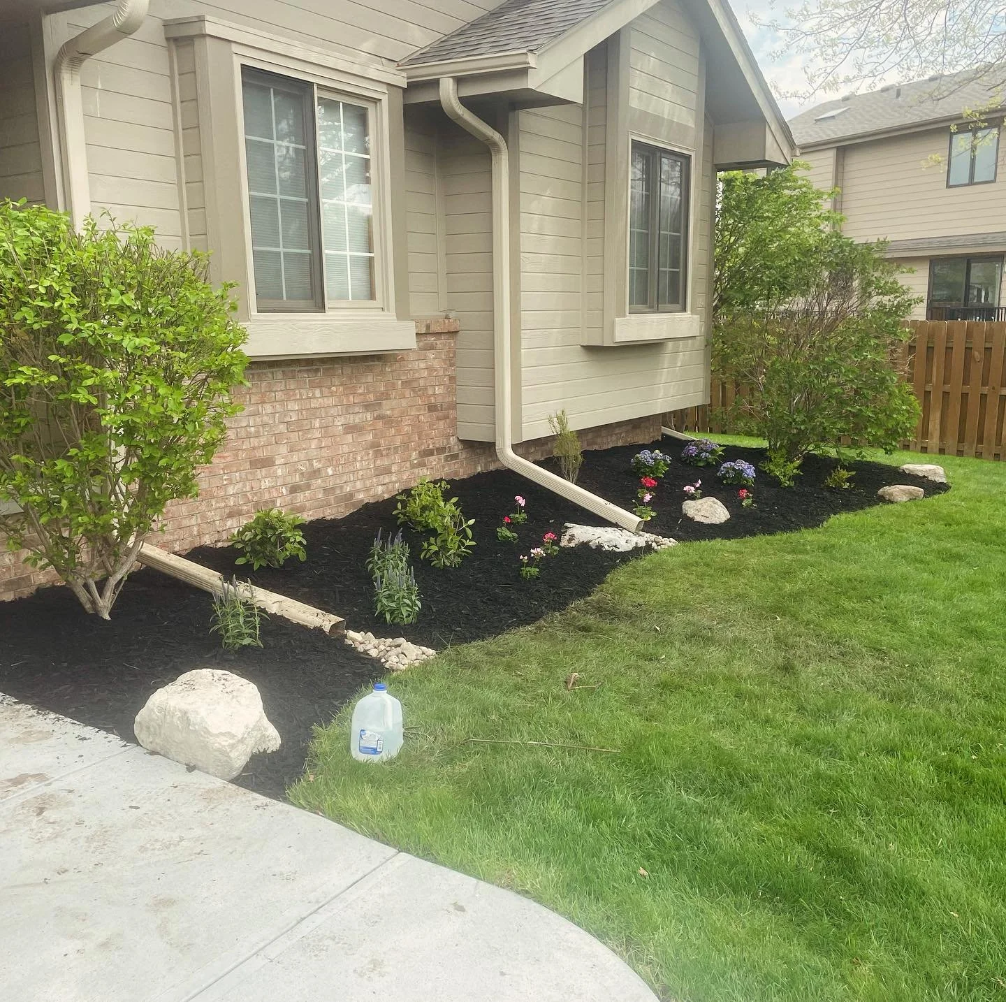 Landscaped front yard with mulched flower beds, small bushes, blooming flowers, rocks, green grass, house with brick and beige siding, white window frames, and gutters.