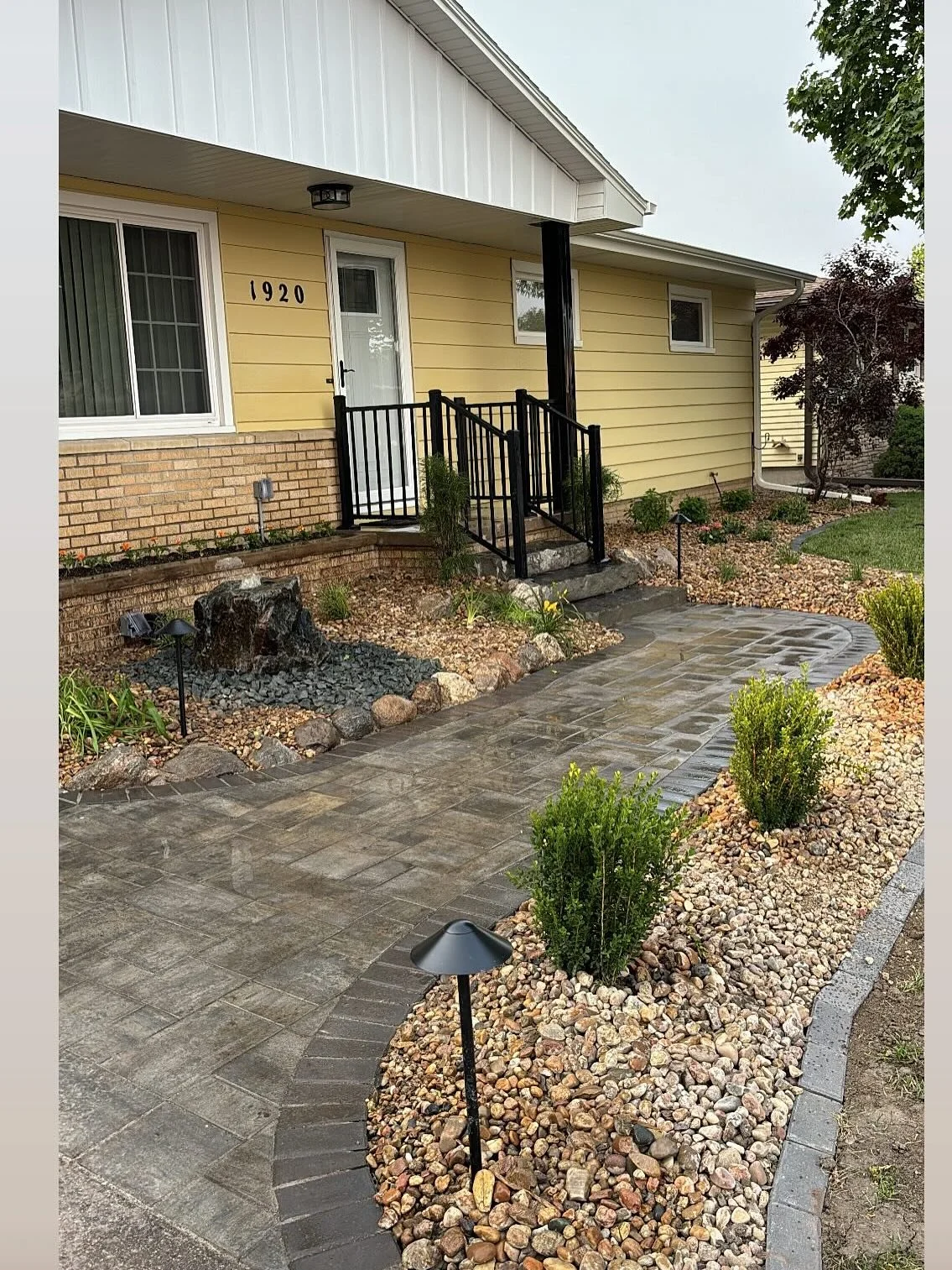 Front yard with a paved walk leading to a yellow house with a small porch and black railing, landscaped with rocks, small shrubs, and garden lights.