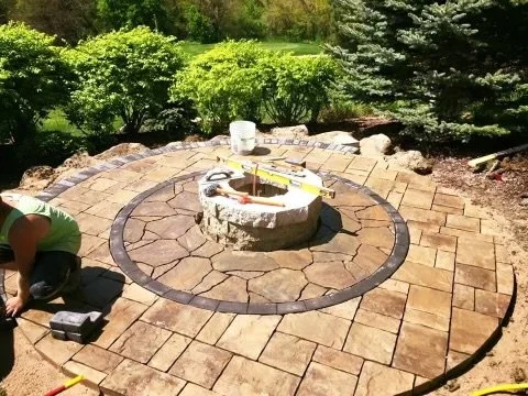 A circular brick patio under construction with a central fire pit, partially built, surrounded by paving stones and garden greenery.
