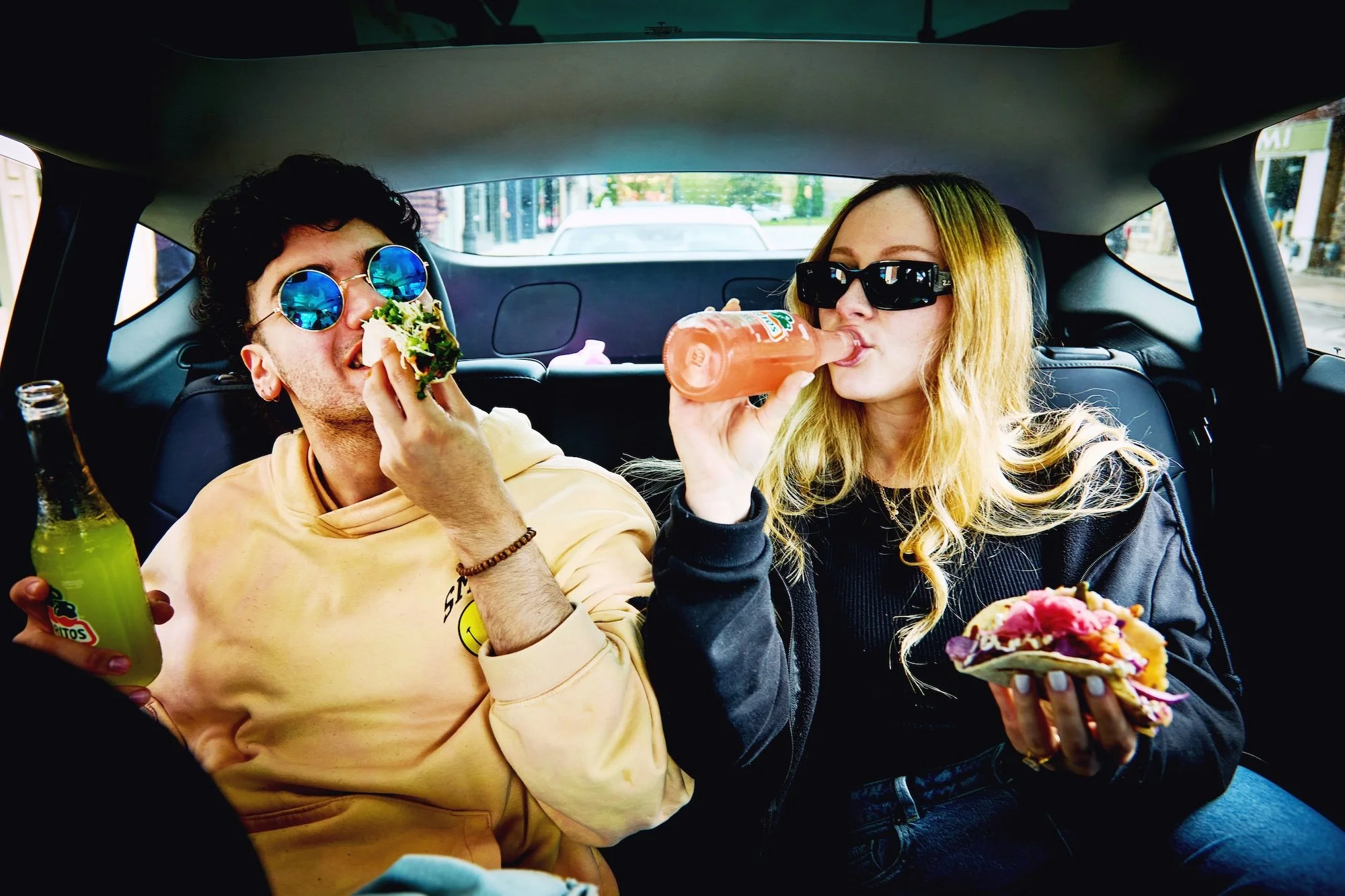 Two young adults sitting in the back of a car, eating tacos and drinking soft drinks. Both wear sunglasses, and the man has curly hair and blue lenses, while the woman has long blonde hair and dark sunglasses.