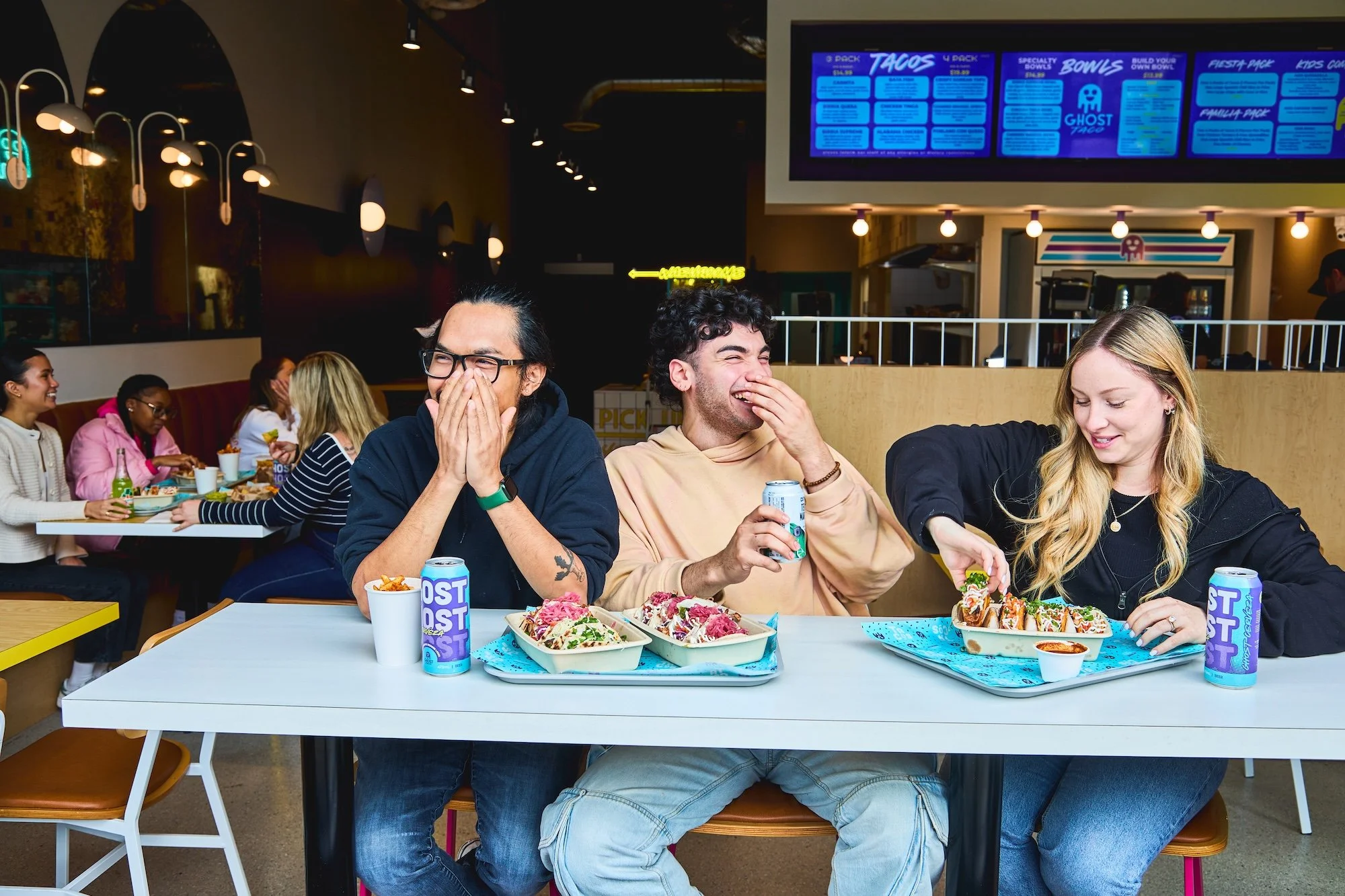 Group of friends laughing and eating tacos at a Ghost Taco restaurant with a digital menu board overhead.
