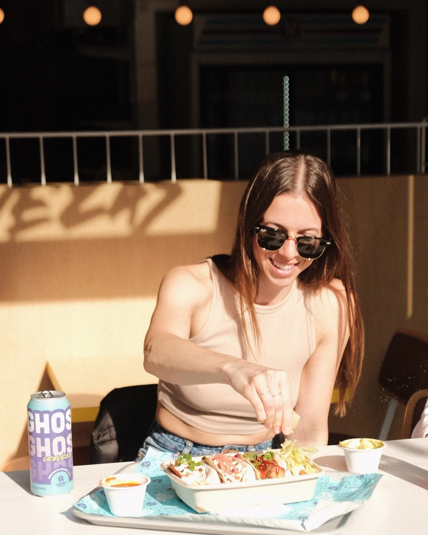 A woman with long brown hair, wearing sunglasses and a beige sleeveless top, is smiling while adding toppings to a tray of tacos at a table. There is a can of Ghost beer and small bowls of sauces or dips on the table.