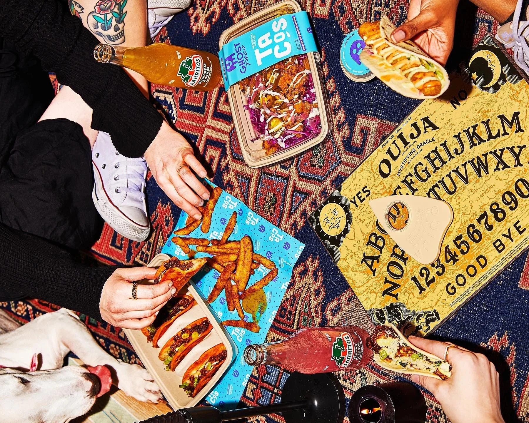 Food and drinks on a patterned table with a Ouija board. There are Ghost Taco tacos and fries with people holding and eating the food. A bottle of Jarritos soda and a salad in a plastic container are visible, along with a dog at the bottom left.