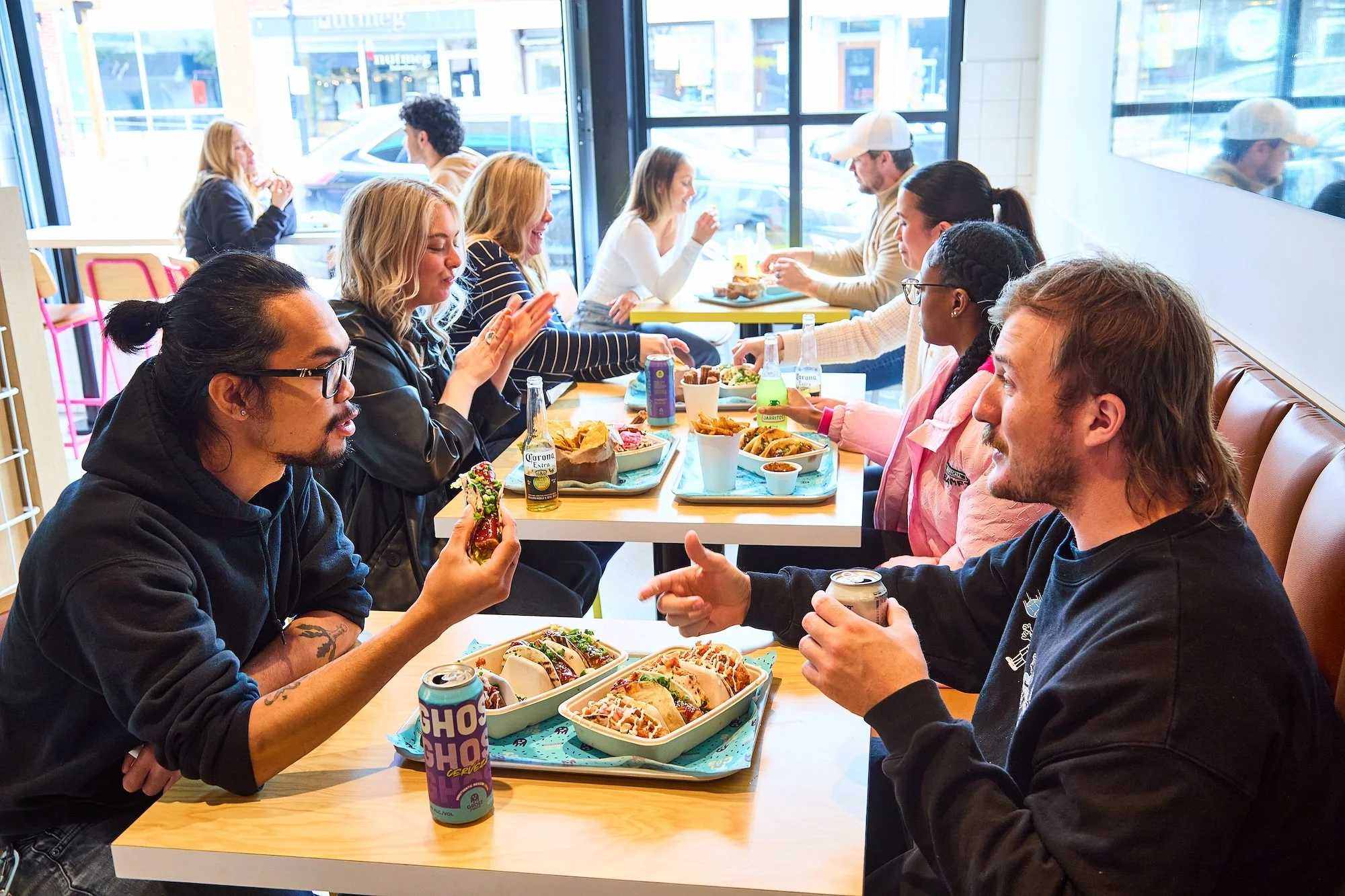 People dining at a Ghost Taco restaurant enjoying tacos, sandwiches, and drinks during daytime.