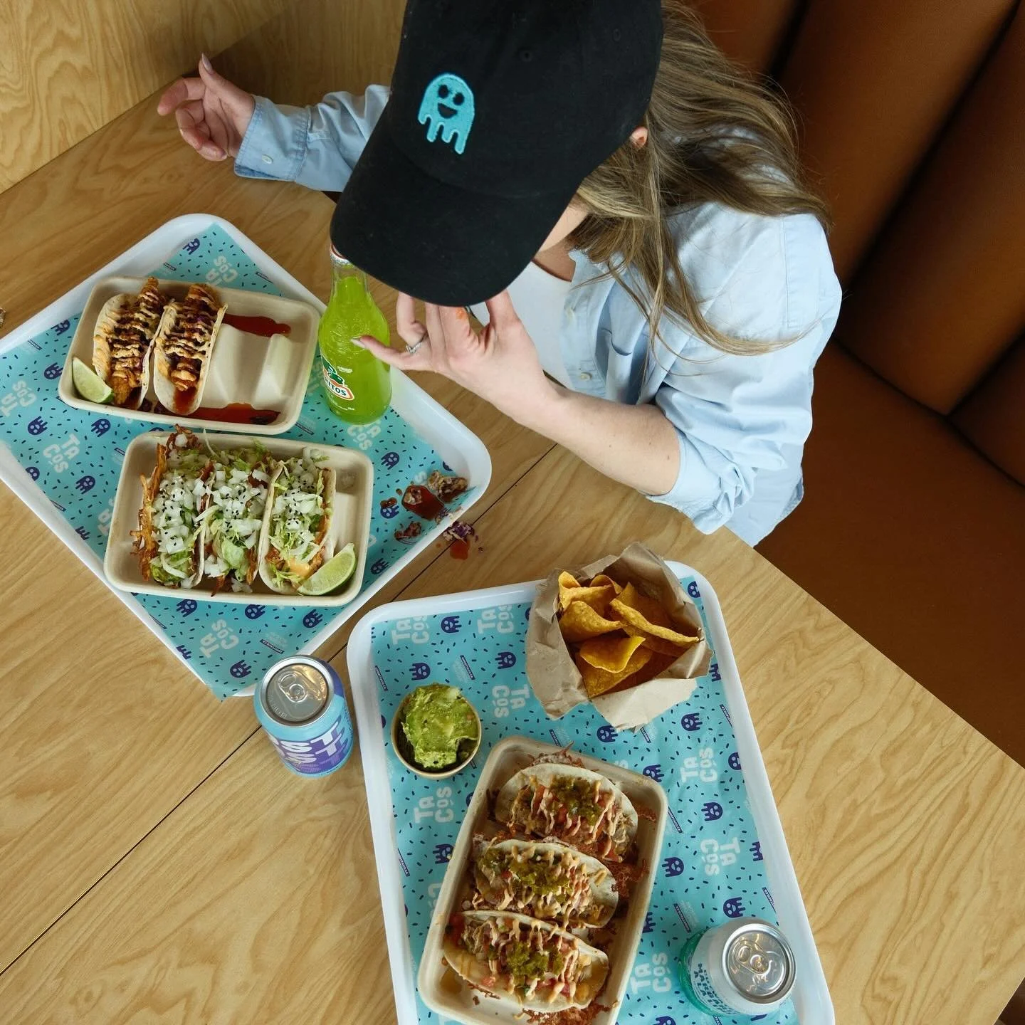 A person wearing a black cap with a blue alien graphic eating tacos at a table with trays of Mexican food, including tacos, chips, and drinks. The setting appears to be a restaurant or fast food place.