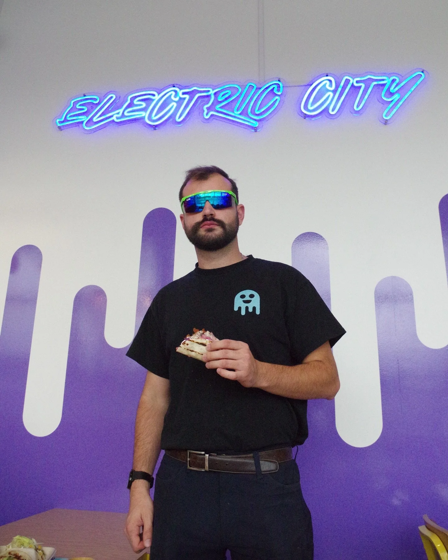 A man with a beard and wearing sunglasses, a black T-shirt with a blue ghost logo, holding a piece of cake, standing in front of a wall with a neon sign that reads 'ELECTRIC CITY'.