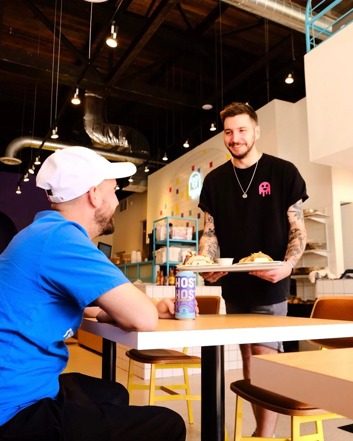A smiling man with tattoos on his arms serving food to another man seated at a table in a Ghost Taco franchise with colorful decor.