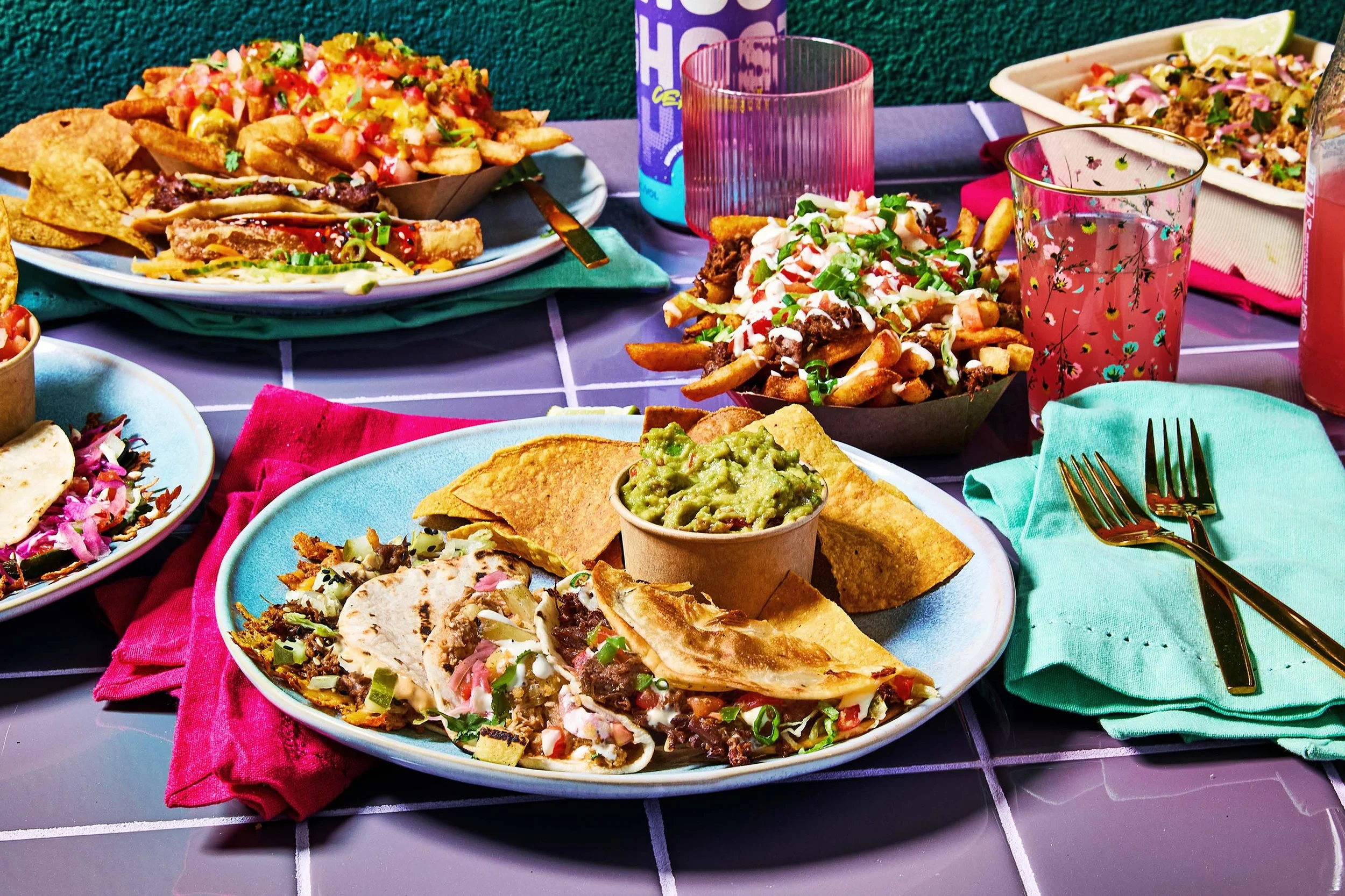 Colorful outdoor table setting with plates of tacos, nachos, and salads, along with pink glasses and summer drinks on a purple tiled surface.