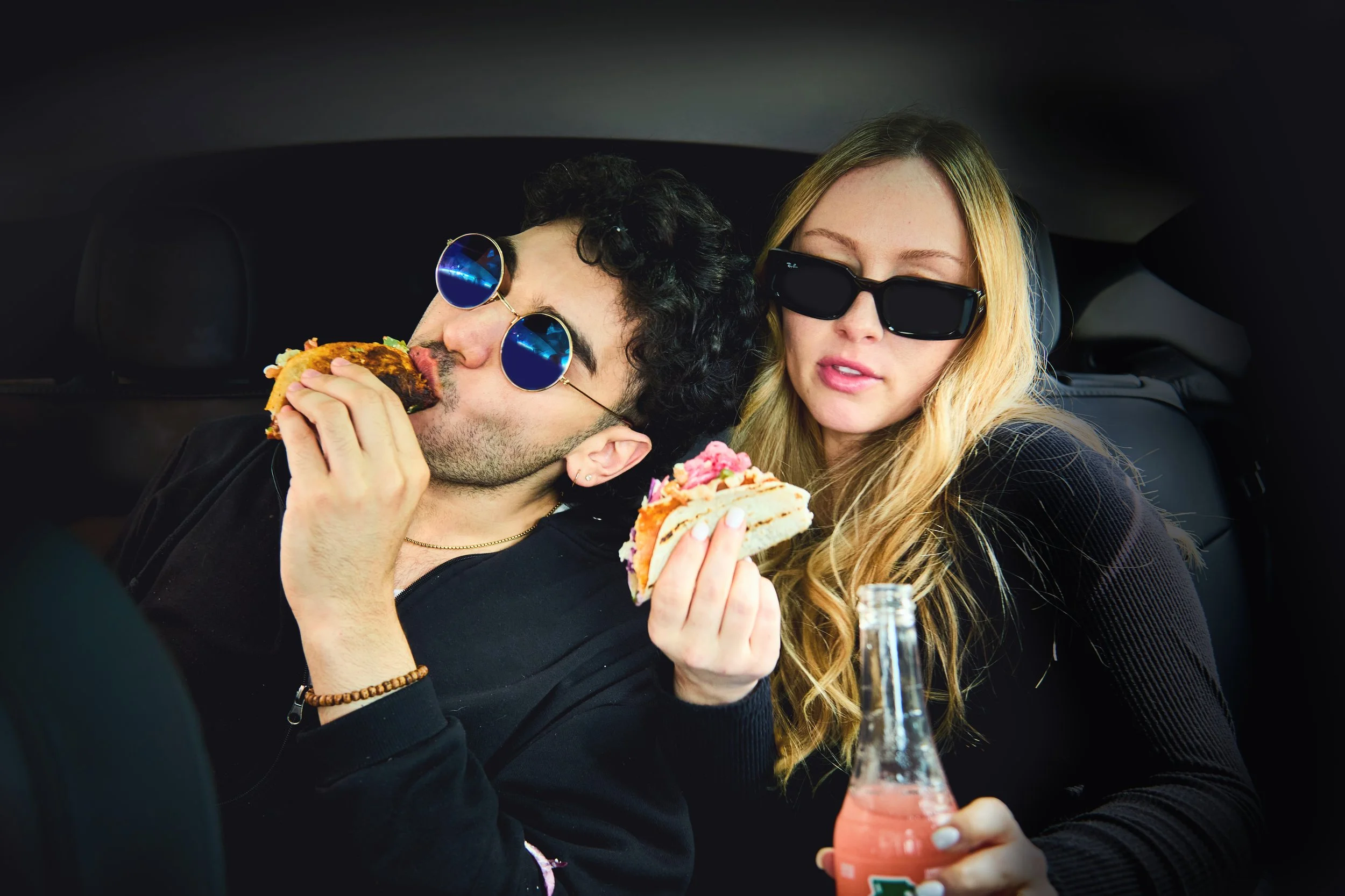 A young man and woman wearing sunglasses sitting in a car, sharing tacos and a soda.