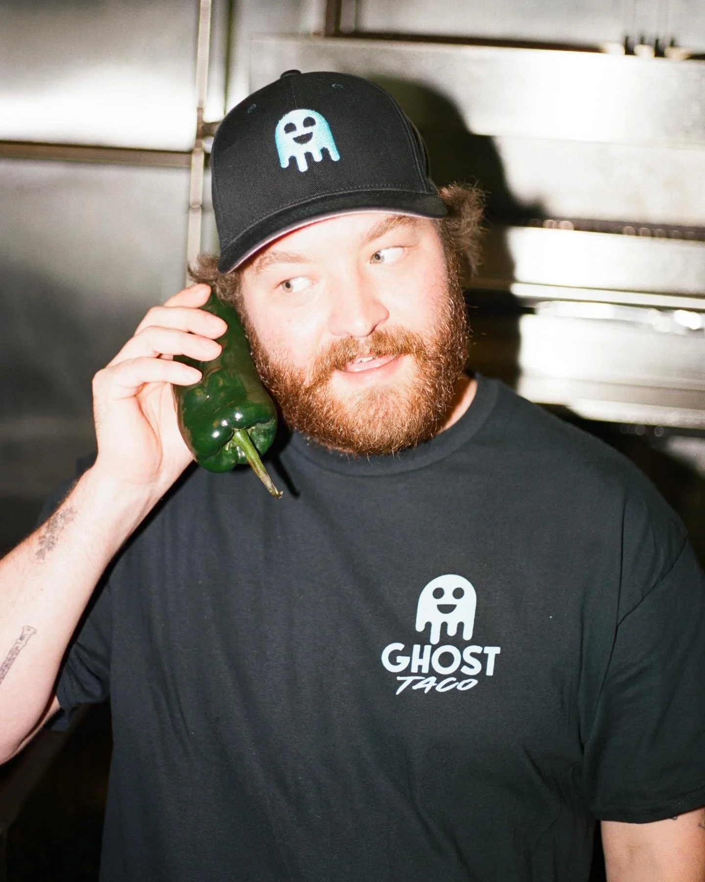 A man with a red beard and brown hair, wearing a black cap with a blue ghost logo and a black T-shirt with the Ghost Taco logo, holding a green bell pepper to his ear as if it were a phone, in a kitchen setting.