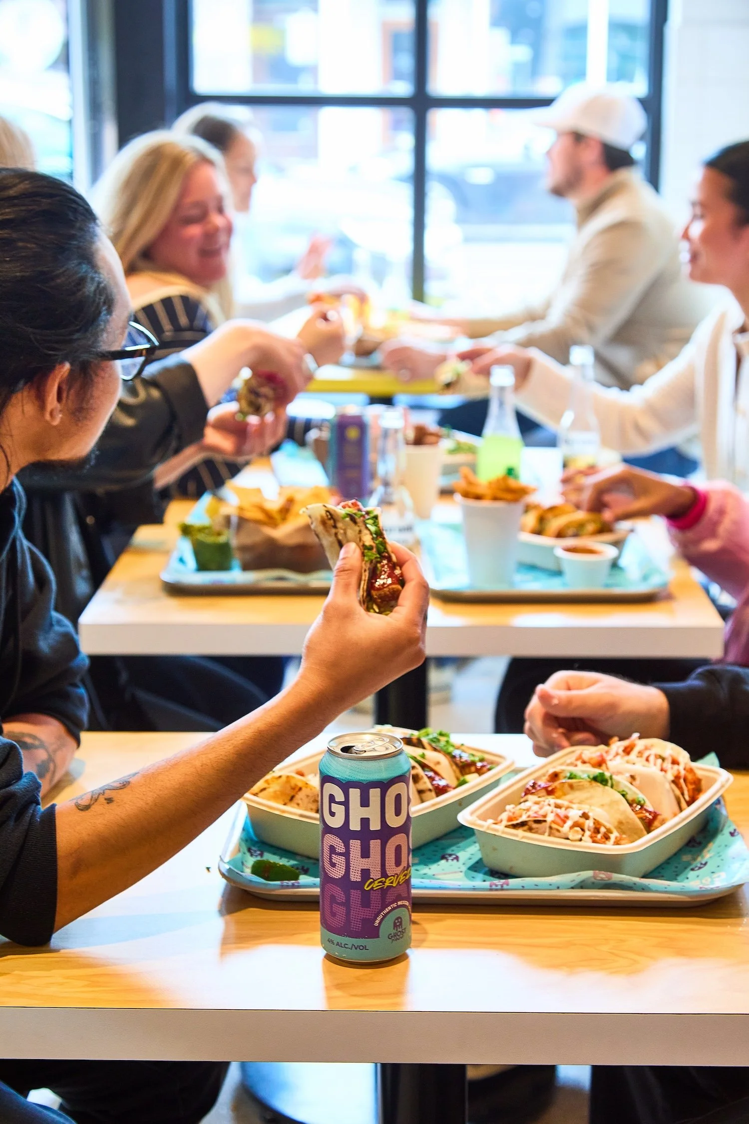 People enjoying a meal at a Ghost Taco restaurant, with tacos, burritos, and a GHOST cervesa beverage on the table.