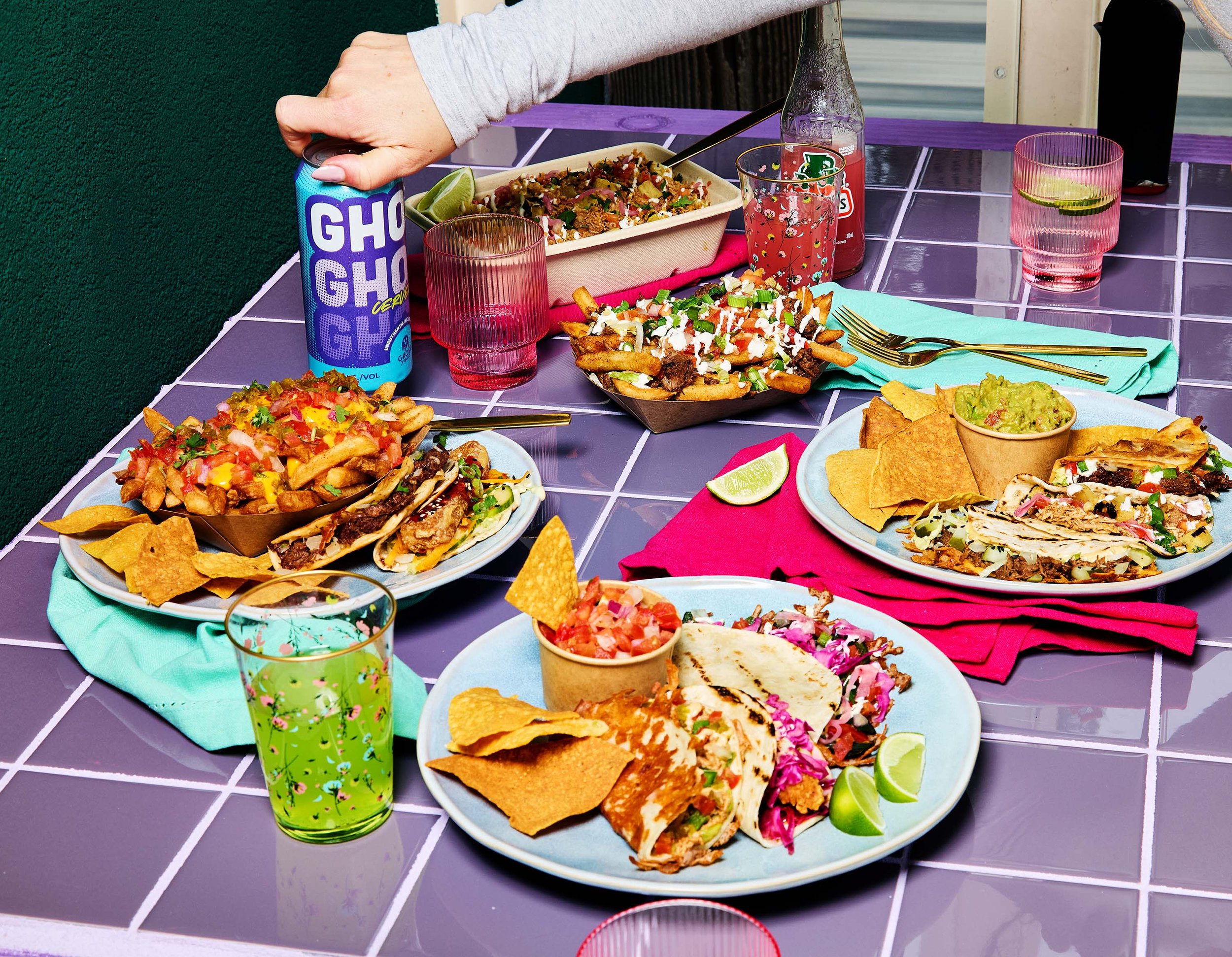 Colorful spread of Ghost Taco Mexican foods on a purple-tiled table, including tacos, chips, guacamole, and salads, with colorful glasses and napkins.