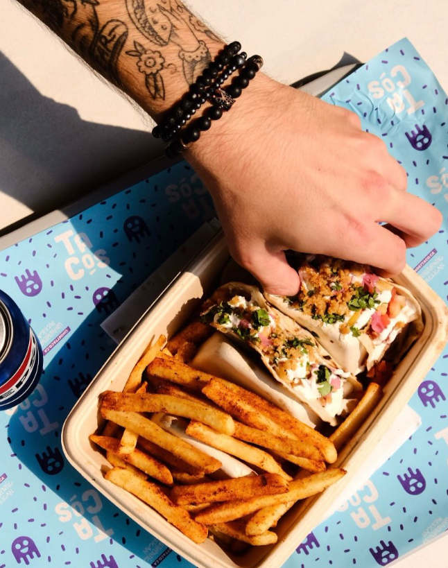 Hand reaching into a takeout container filled with fries and a wrap. The hand has tattoos and is wearing black beaded bracelets. A can of soda is partially visible on a blue tray liner with taco-themed graphics.