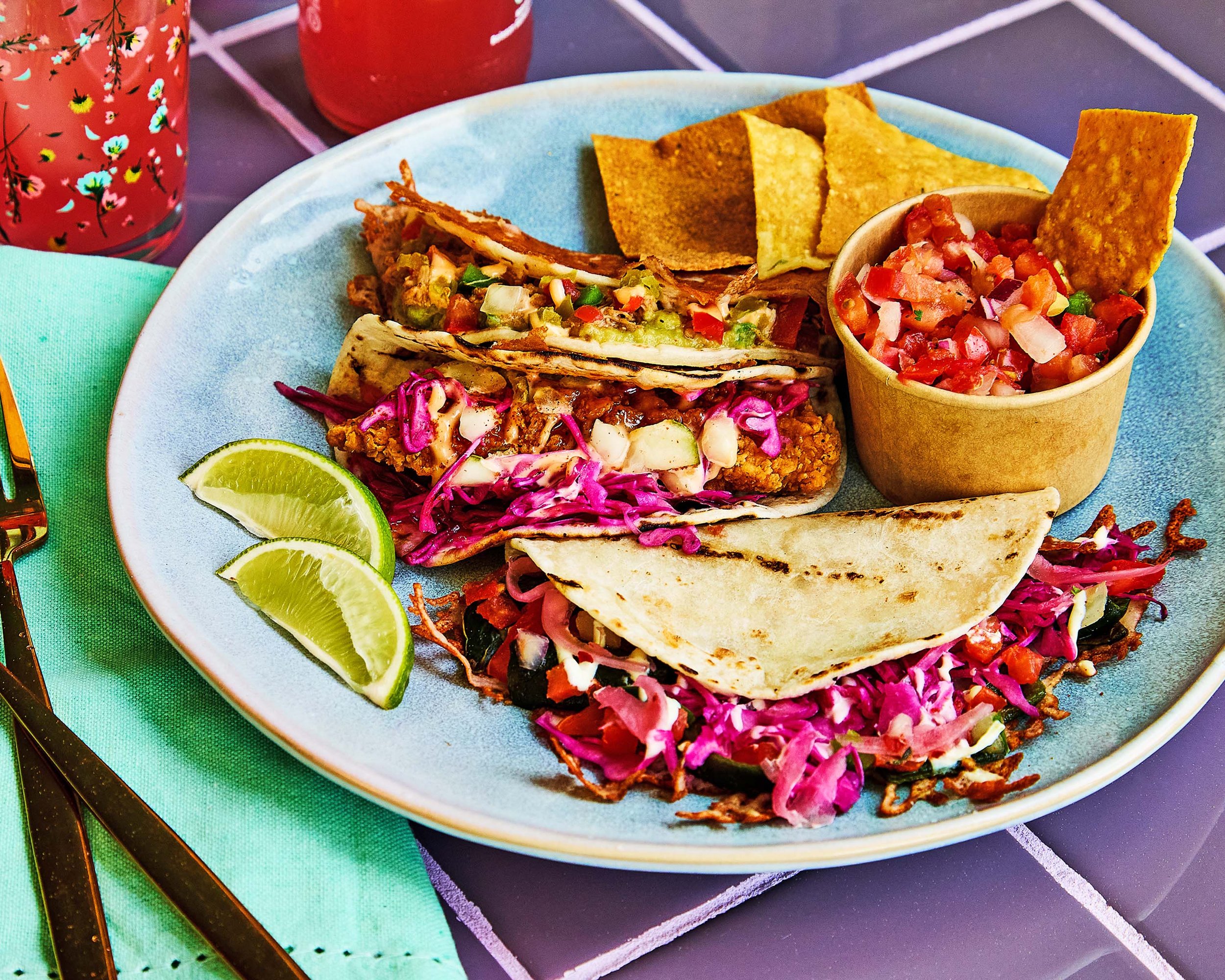 Plate of tacos with lime wedges, served with a side of salsa, tortilla chips, and grilled meat.