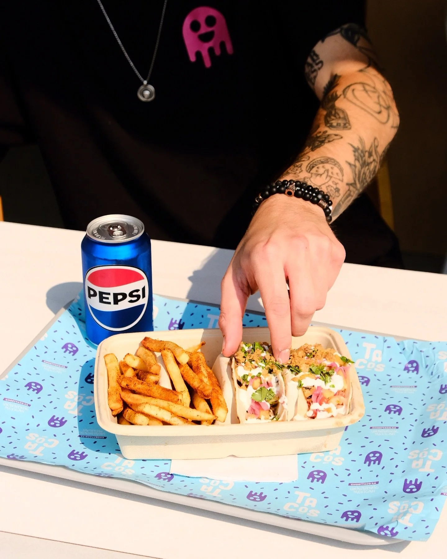 A person with tattoos on their arm, wearing a black T-shirt with pink ghost and necklace, reaching into a cardboard food container with fries and tacos, on a blue Ghost Taco dispenser tray, with a can of Pepsi on the side.