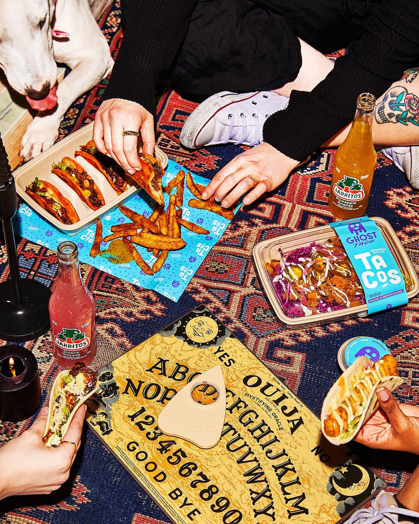 People having a picnic on a colorful rug with tacos, fries, a salad, drinks, a ouija board, and a tarot card.