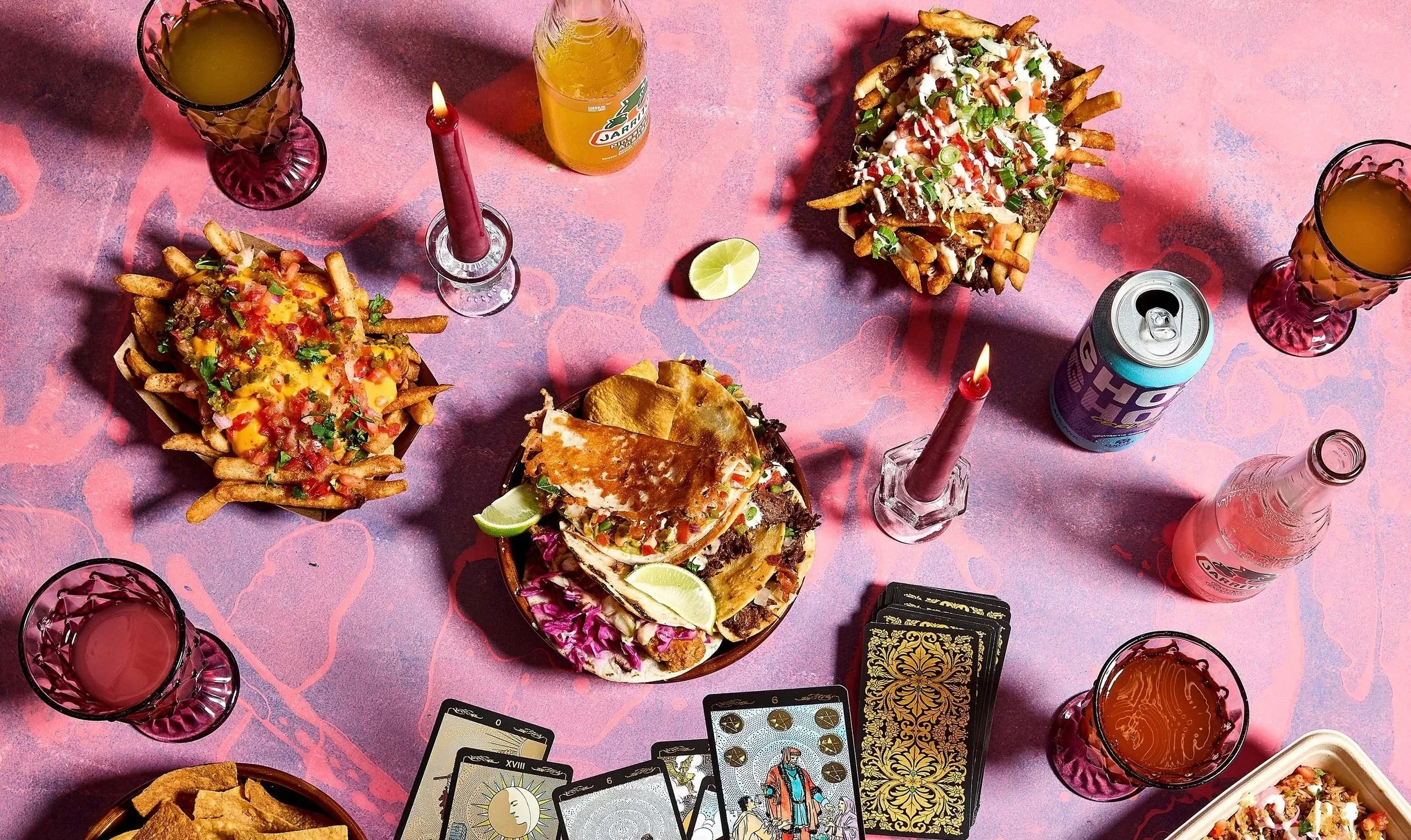 Table with various Mexican foods including nachos, tacos, and fries, along with drinks, candles, tarot cards, and a lime wedge on a pink tablecloth.