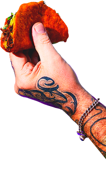 A hand with tattoos and a bracelet holding a taco with meat, vegetables, and sauce.