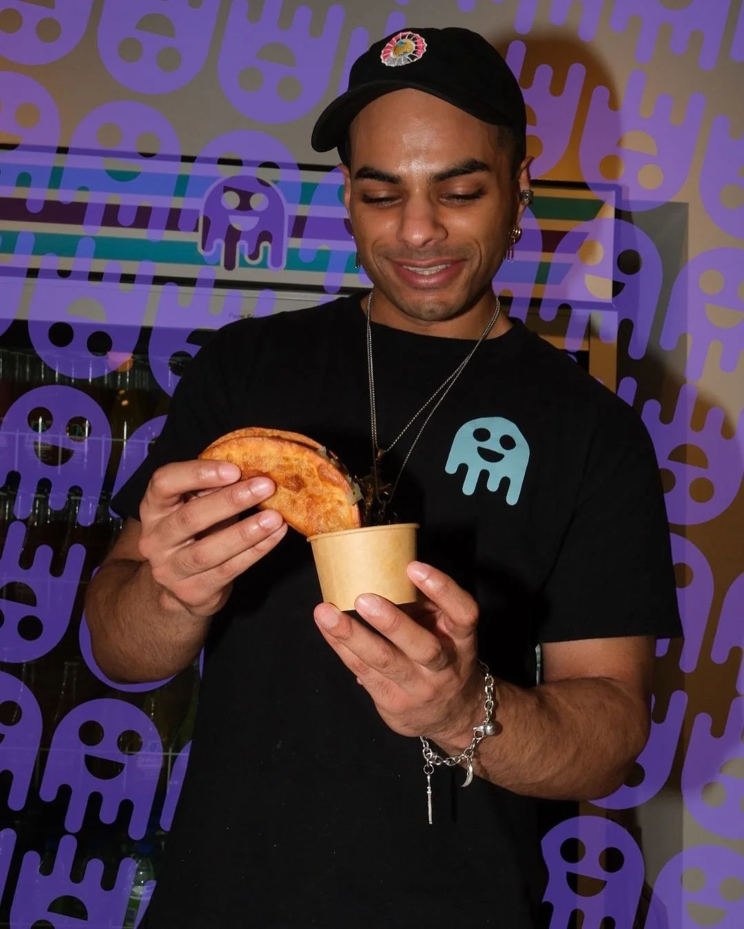 A man wearing a black shirt and cap, holding a taco in his right hand and a small paper cup in his left hand, smiling at the food.