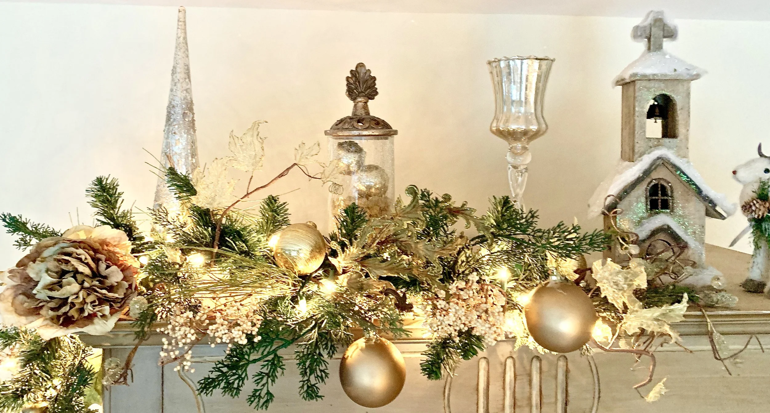 Decorative Christmas display with greenery, gold ornaments, a pine cone, and ceramic village pieces on a mantelpiece.