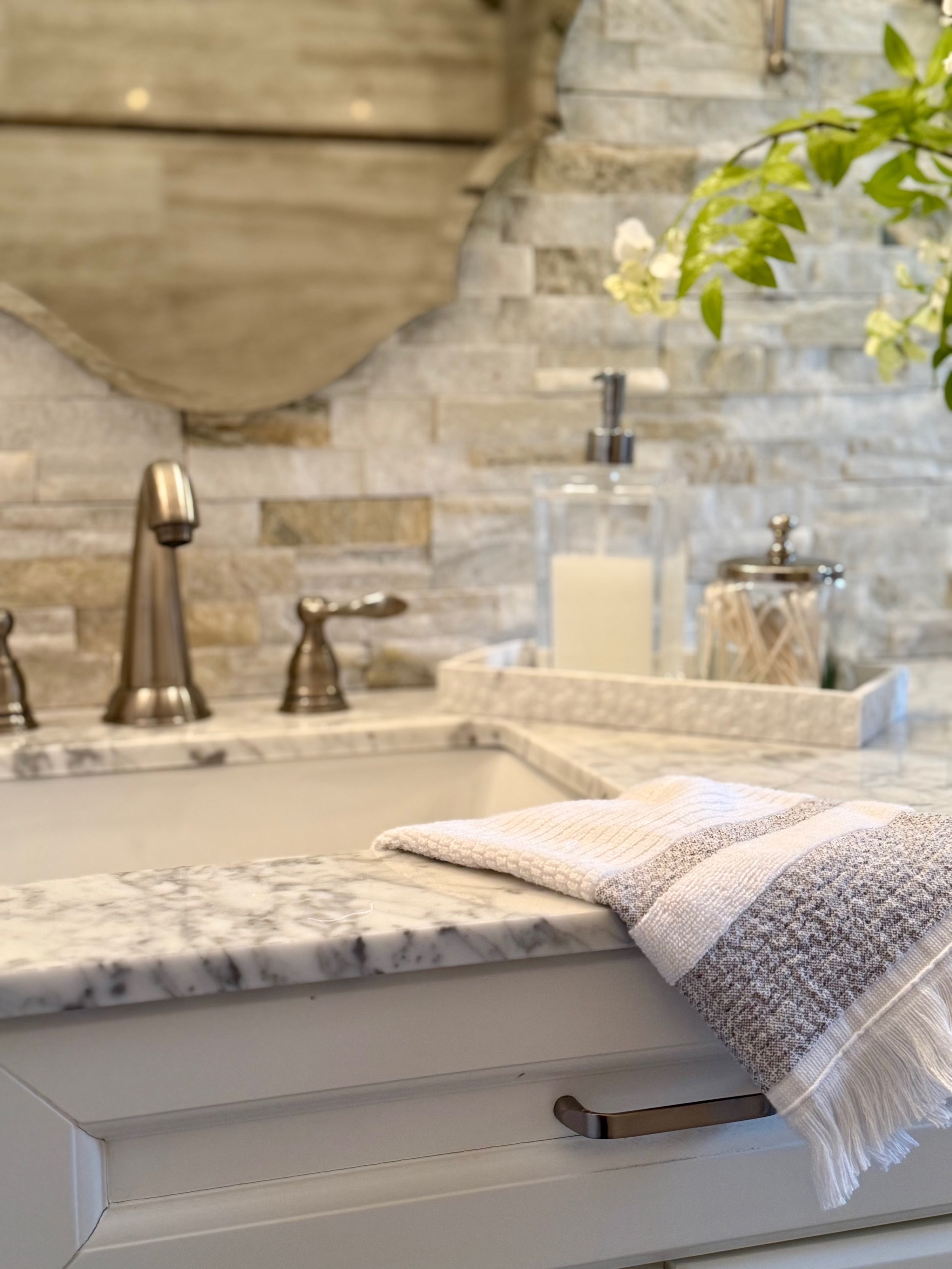 grey and white marble bathroom vanity with a grey and white towel, a marble tray holding a clear and silver glass soap dispenser and glass and silver container, brushed silver faucet, green and white florals against a grey ledger stone backsplash