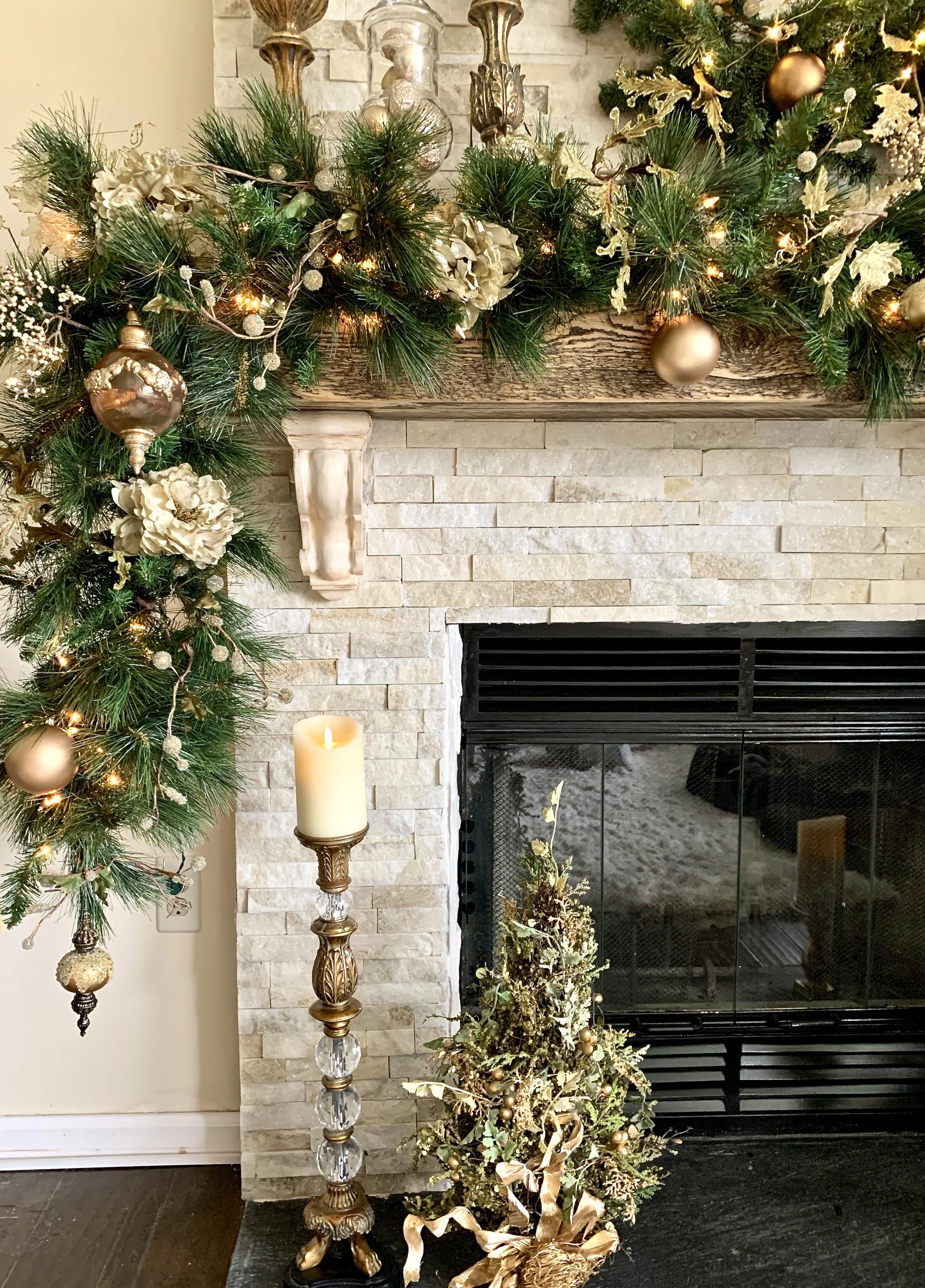 Decorated fireplace mantel with greenery, gold ornaments, and lights, next to a small gold and green Christmas tree and a tall candle on a decorative candlestick.