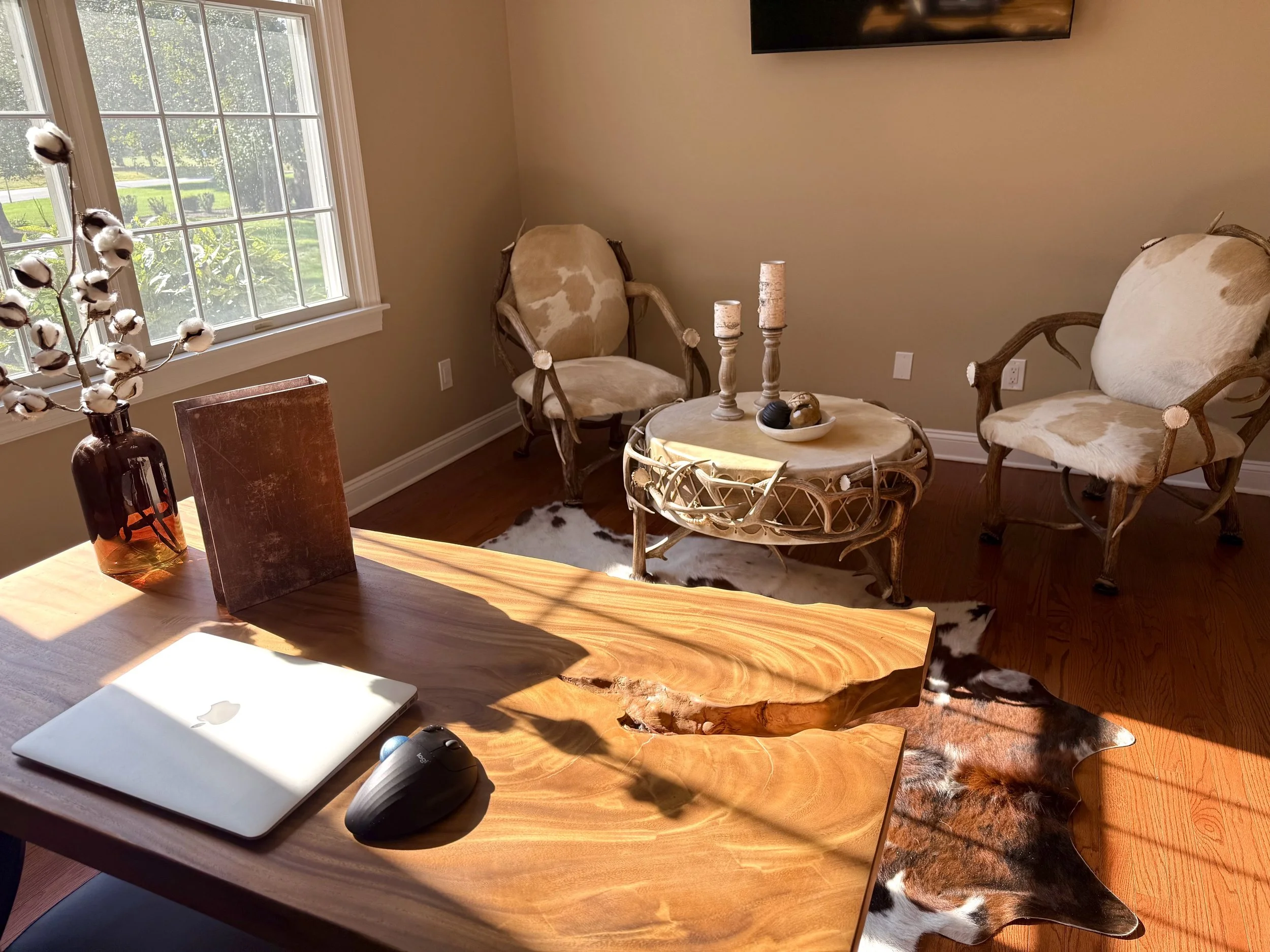 Living room with two cowhide rugs, a wooden coffee table with candles, two beige cowhide chairs, and a table with a cotton branch in a vase near a large window letting in sunlight.
