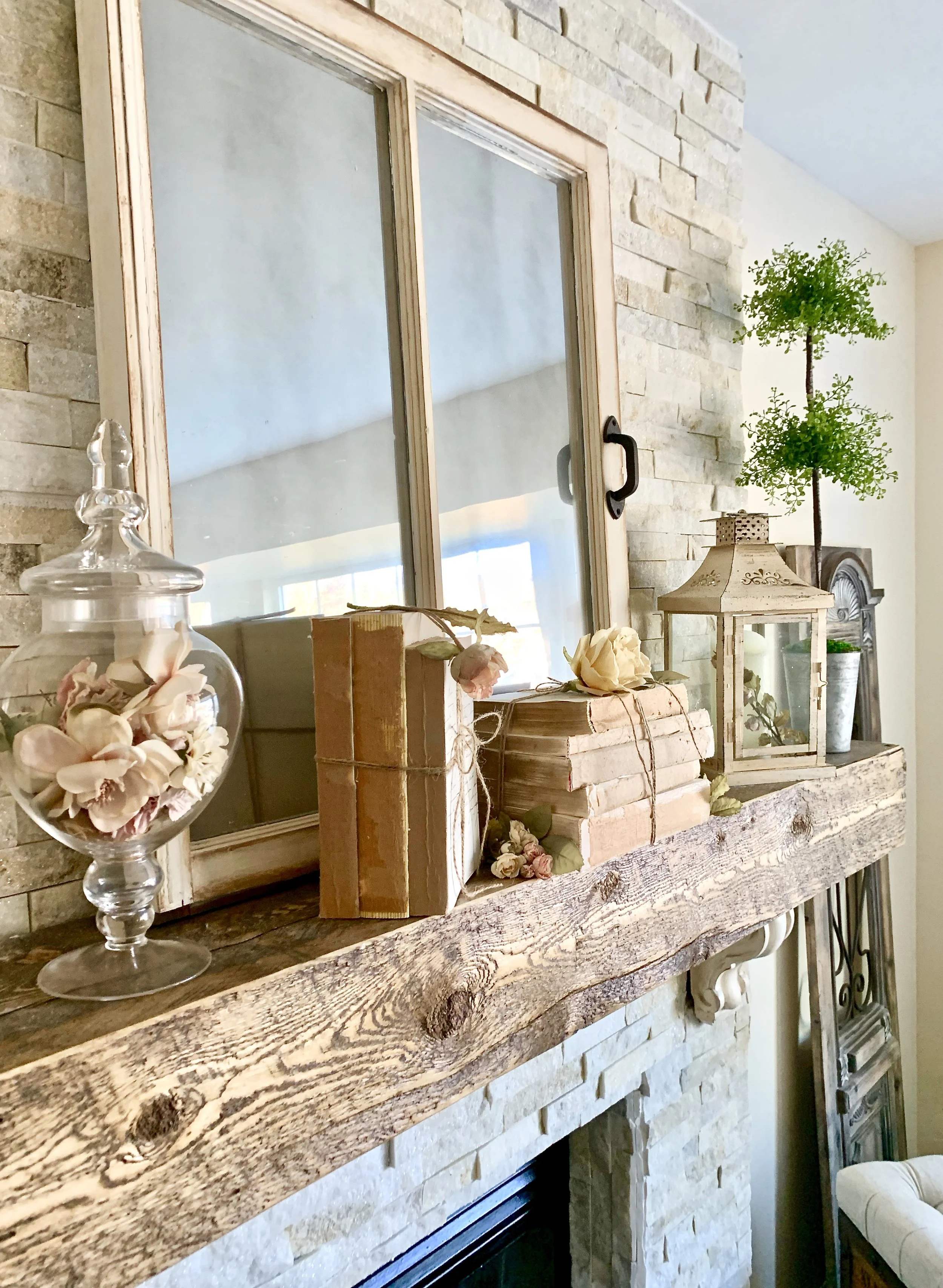 Decorative style living room featuring a wooden mantel with vintage books, flowers, lantern, and a plant, framed mirror, and stone wall.