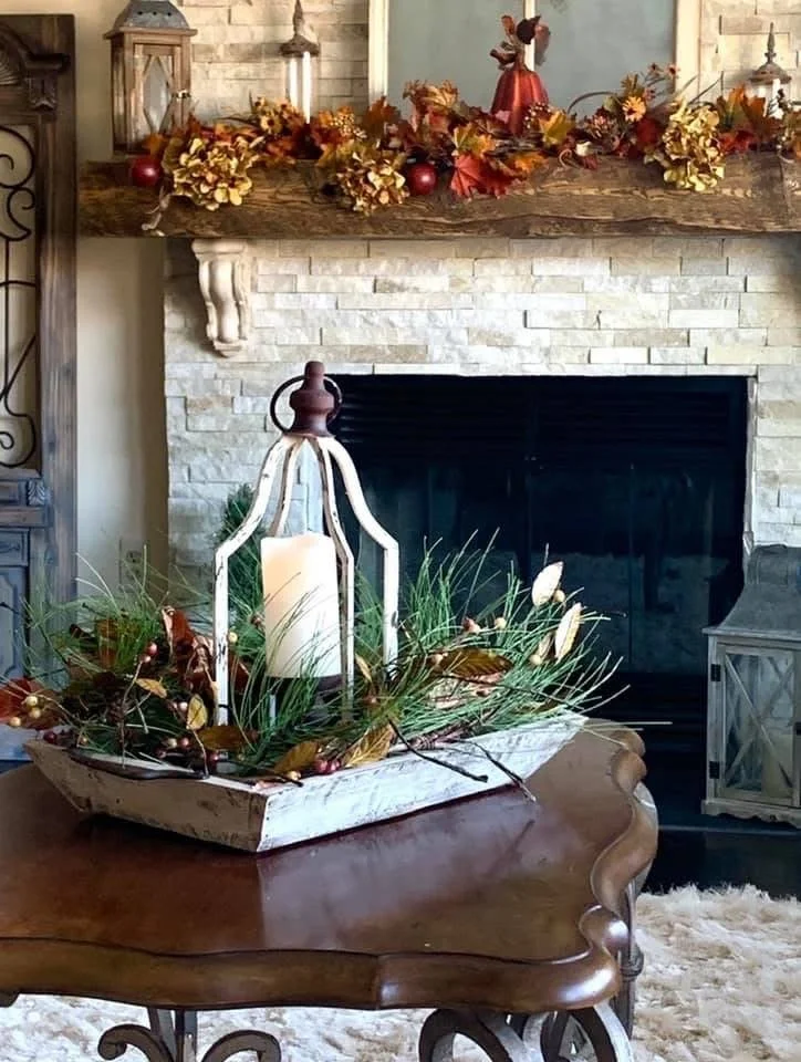 Decorative autumn holiday scene in a living room featuring a wooden table with a centerpiece of green foliage and a lantern with a candle, a fireplace with a stone surround, and a mantel decorated with fall leaves, artificial fruits, and a small ange