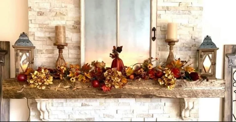 Decorative fall centerpiece on a rustic wooden mantle with candles, lanterns, and autumn leaves.