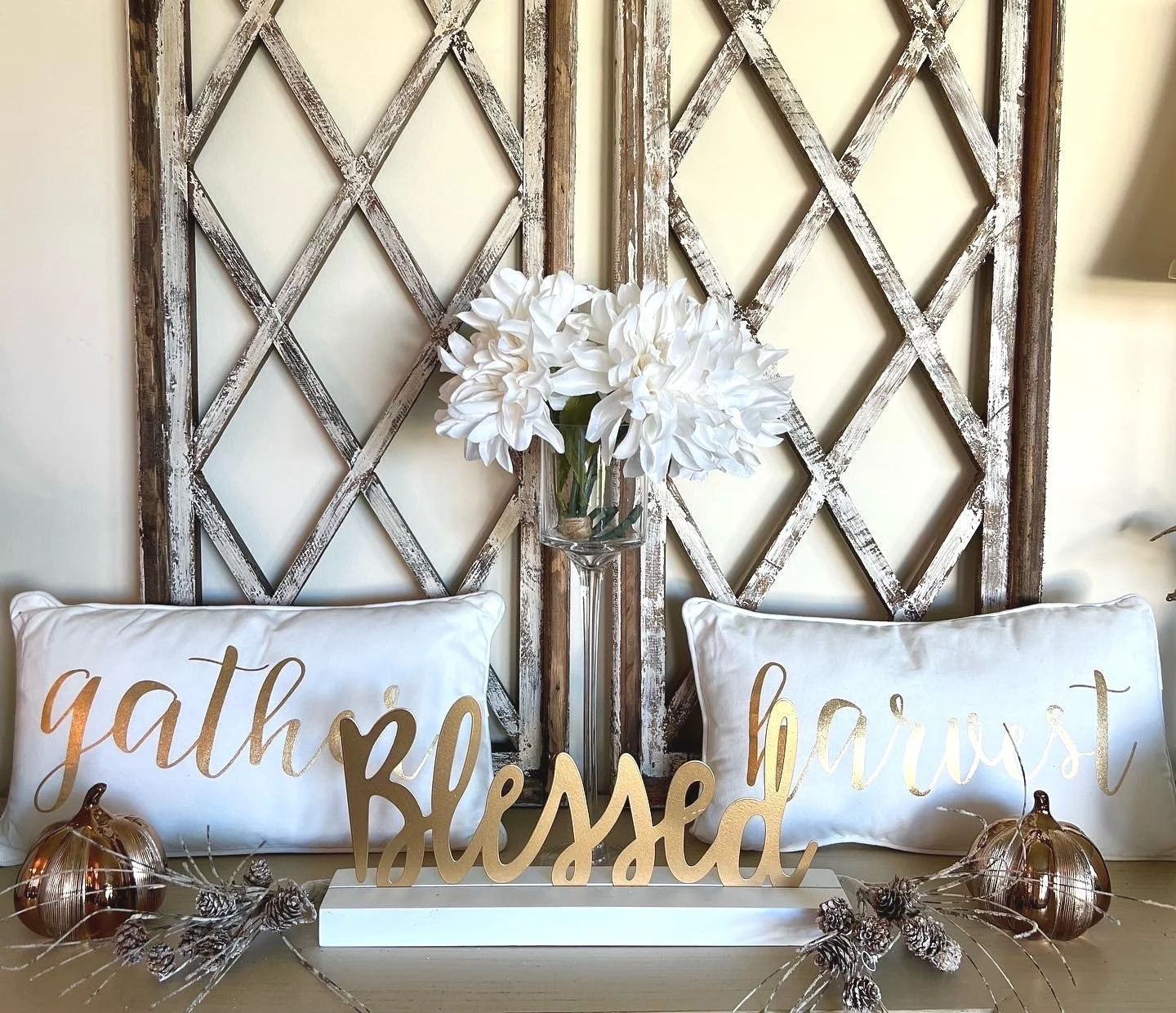 Decorative home display with white pillows that say 'gather' and 'happiest', a tall glass vase with white flowers, gold and copper pumpkins, pinecones, and a wooden sign that says 'blessed', all on a table against a rustic whitewashed wooden backdrop