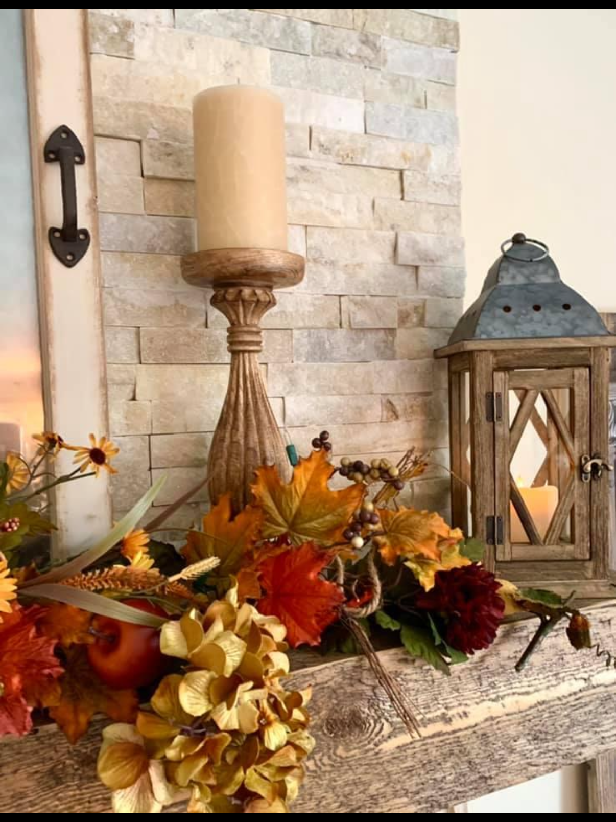 Autumn-themed decorative arrangement featuring a wooden table with fall leaves, berries, flowers, and pomegranates, alongside a tall beige candle on a carved wooden holder and a rustic lantern with a candle inside, set against a stone wall backdrop.