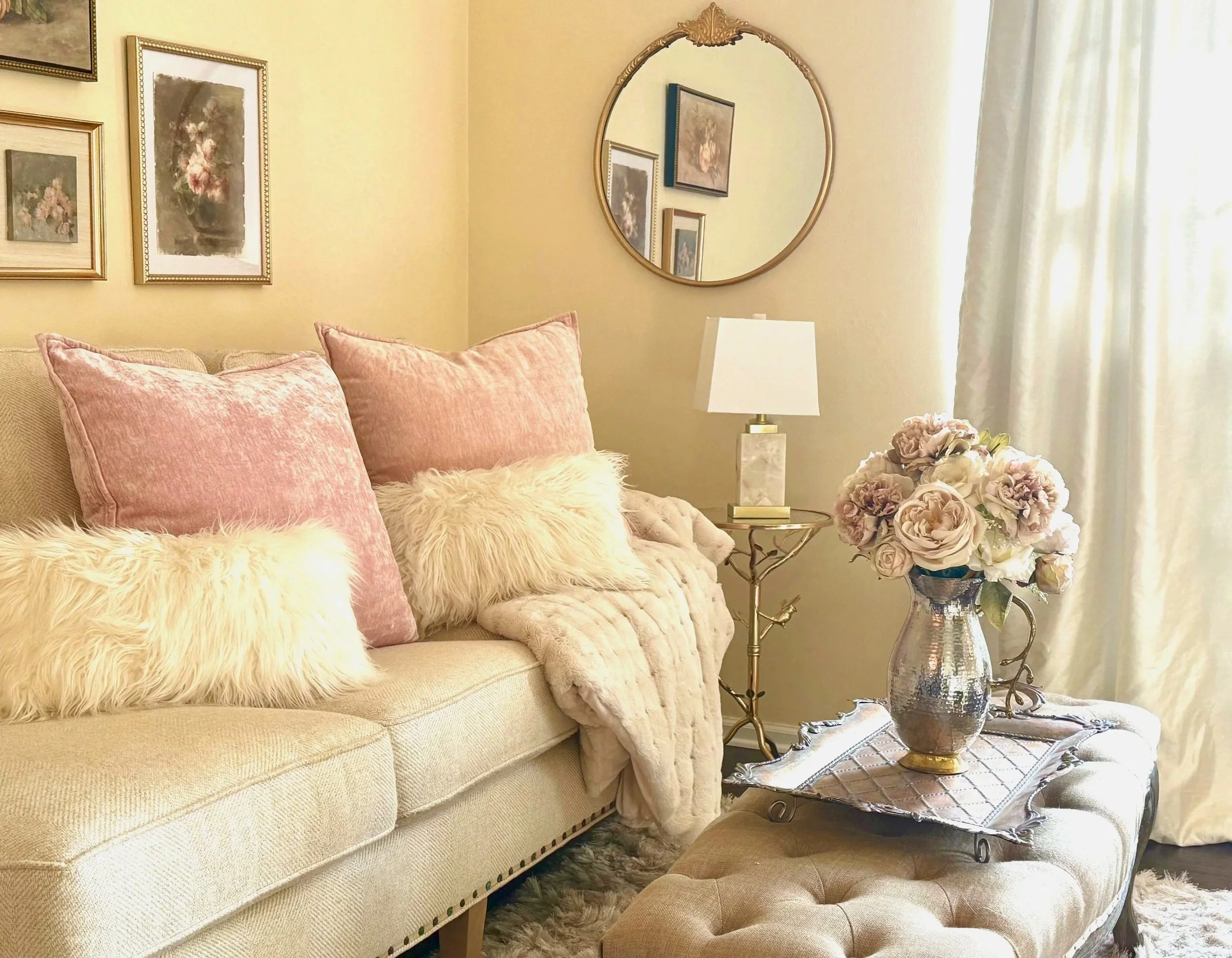 Professionally decorated living room showcasing a neutral sofa with pink and white pillows, a tufted ottoman, layered throw blanket, multiple gold and pink framed artwork, round gold mirror, quartz lamp with white shade, gold and glass side table, si