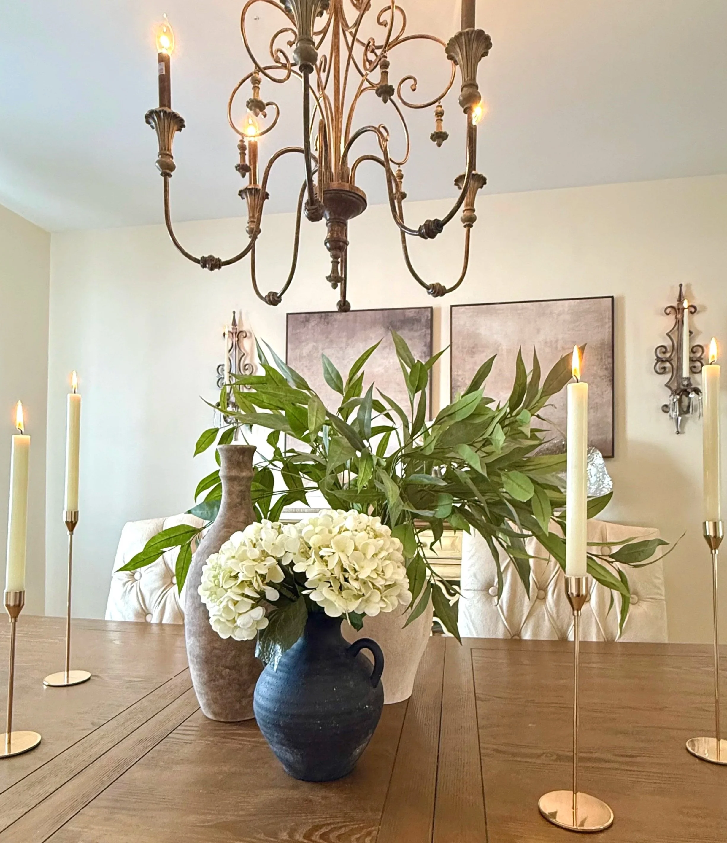 A professionally styled dining room featuring neutral and black ceramic vases filled with lush greenery and white flowers on a wooden dining room table styled with gold pillar candlesticks with an ornate antique-style wrought iron chandelier with can