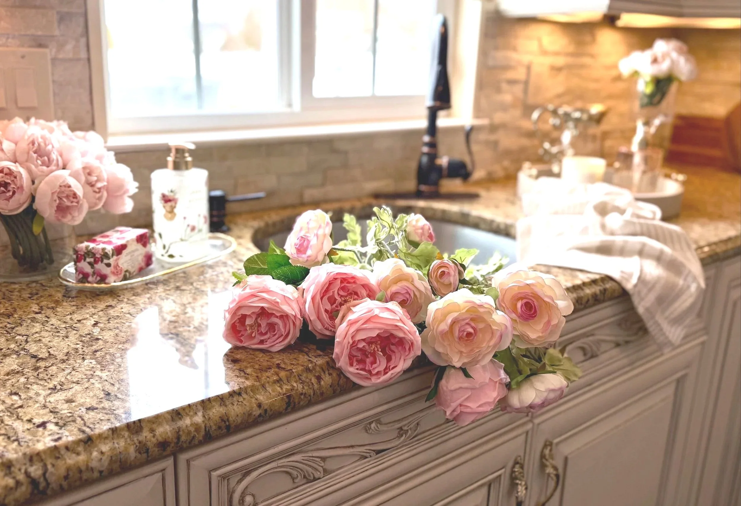 Beautifully styled kitchen counter top with pink flowers cascading from the sink against decorative cabinets. Next to the sink is a gold and glass tray with a pink floral soap dispenser and bars of soap wrapped in a rose pattern paper. A dish towel d