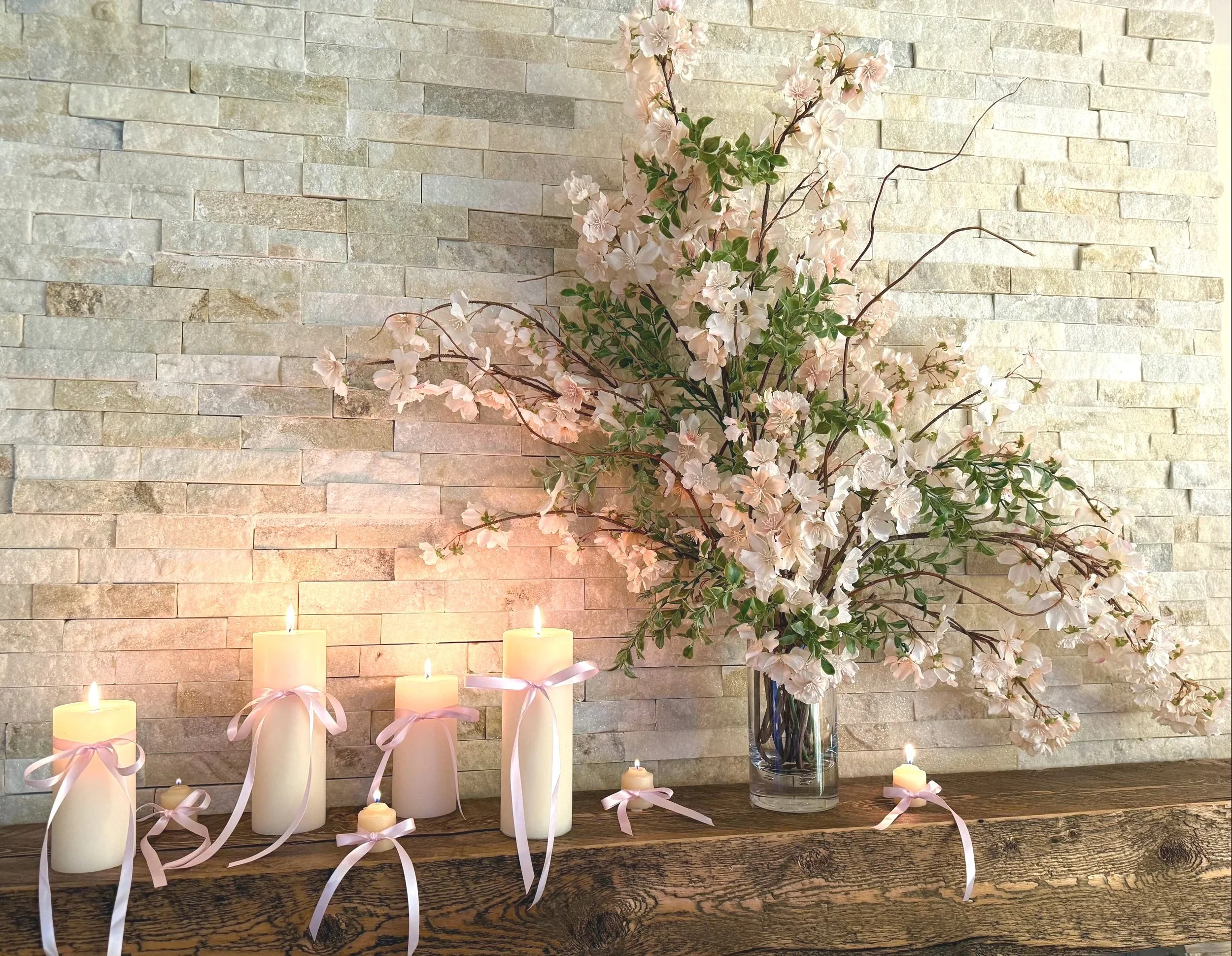 fireplace rustic wooden mantle with tan, beige and white ledger stone surrounding with a glass vase filled with cherry blossoms and 
multiple cream colored candles of various heights all tied with a pink ribbon.