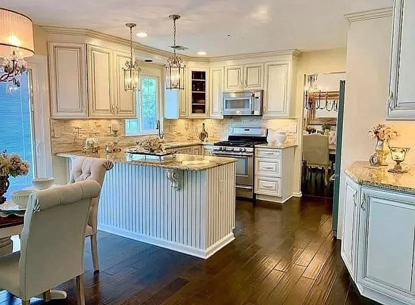 Renovated kitchen with dark hardwood floors and cream distressed cabinets with ornate gold hardware, granite countertops, beautiful pendant lights, bead board island decorated with corbels.