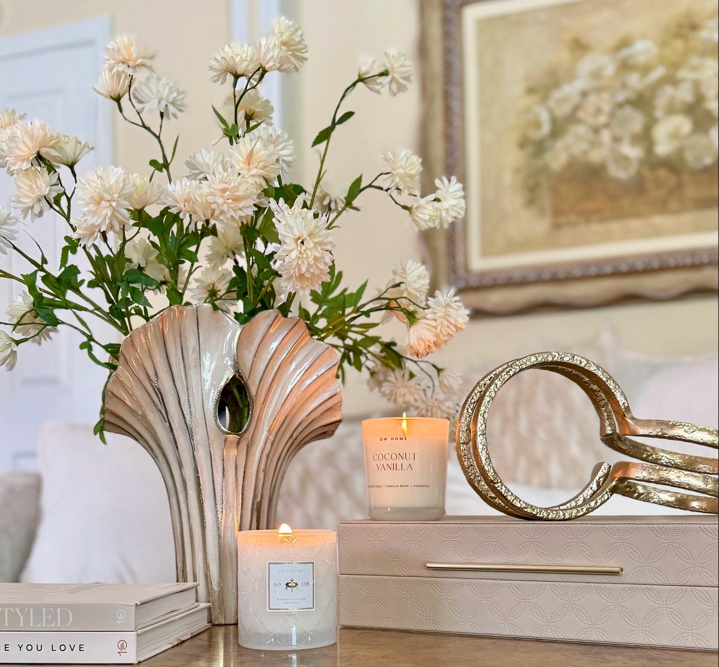coffee table decor with gold and cream colored glass vase filled with bright white flowers paired with decorative books, a tan colored decorative box with a gold sleek handle with a gold abstract accessory that sits on top and lit candles enhancing t