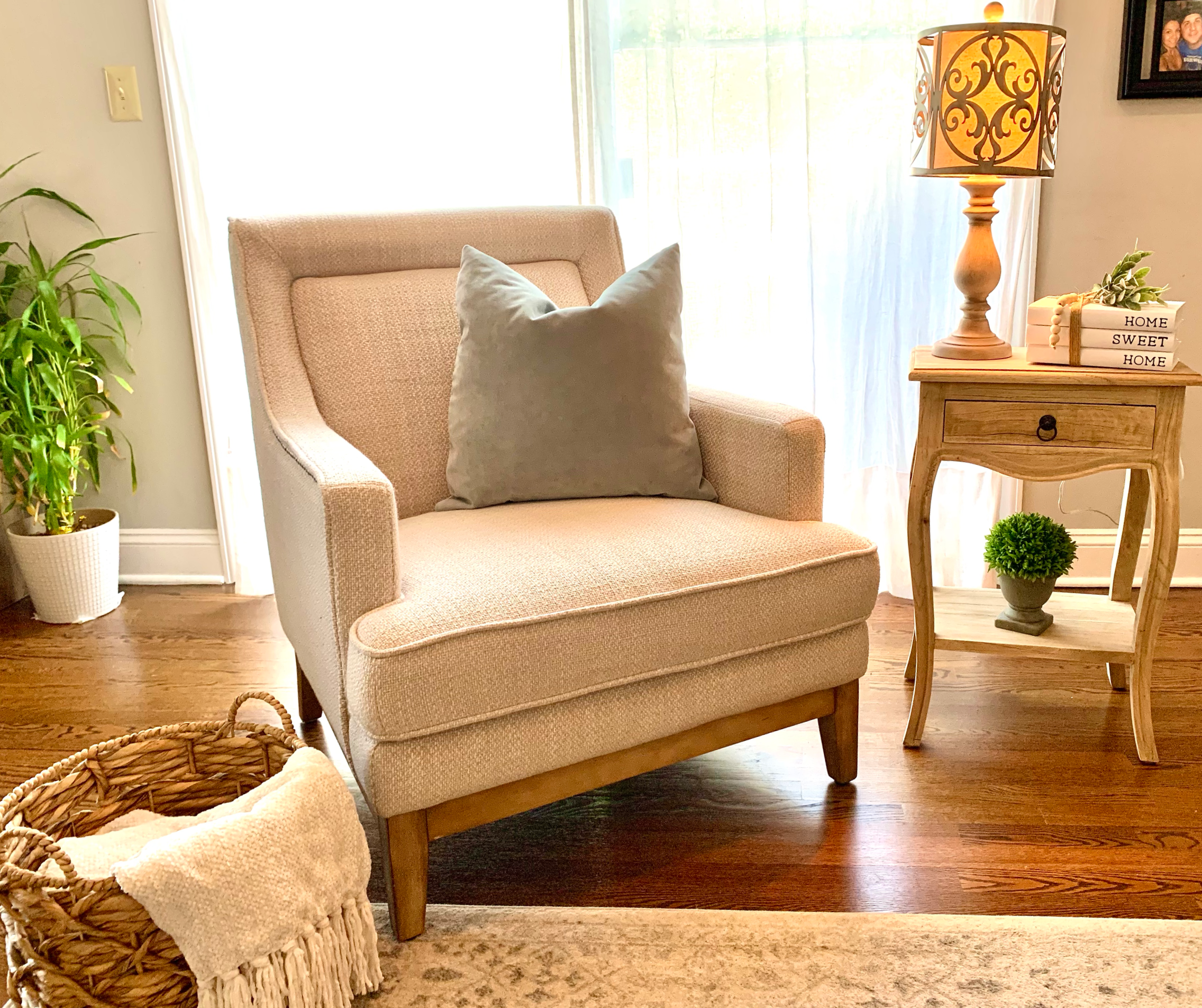 Living room with beige armchair with cushion, side table with decorative items and lamp, potted plant, basket with cozy blanket, hardwood floor, window with sheer curtains.