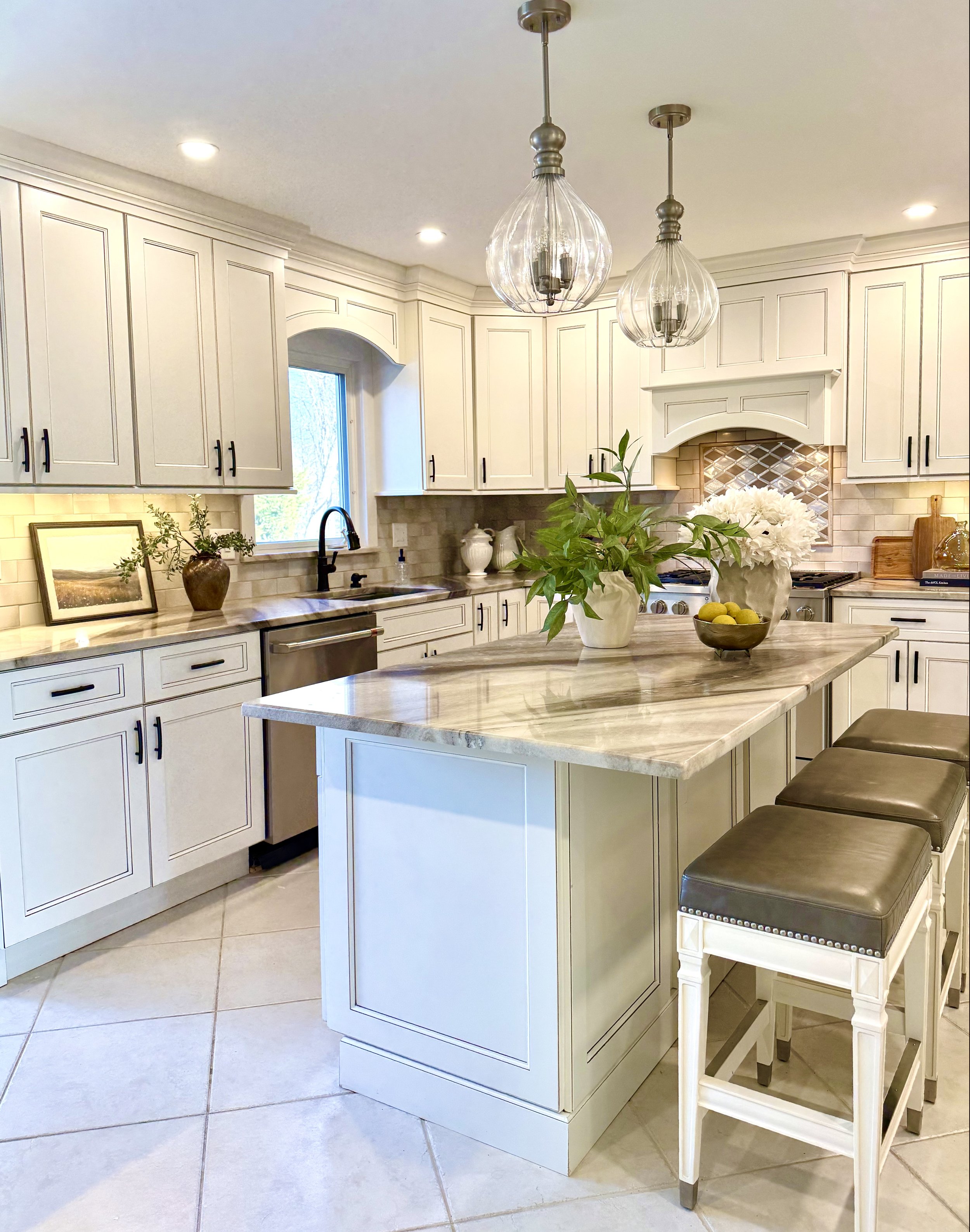 Elegant spacious white kitchen with enlarged countertops, upholstered bar stools, glass pendant lighting, custom cabinetry, decorative greenery, beige, brown and white with black pulls. Designed for a warm and inviting luxury home interior. 