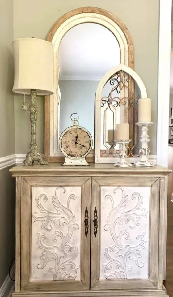 A vintage-style wooden sideboard with intricate carved swirls on the doors, topped with candles, a clock, lampshades, and mirrors in natural wood and white finishes.