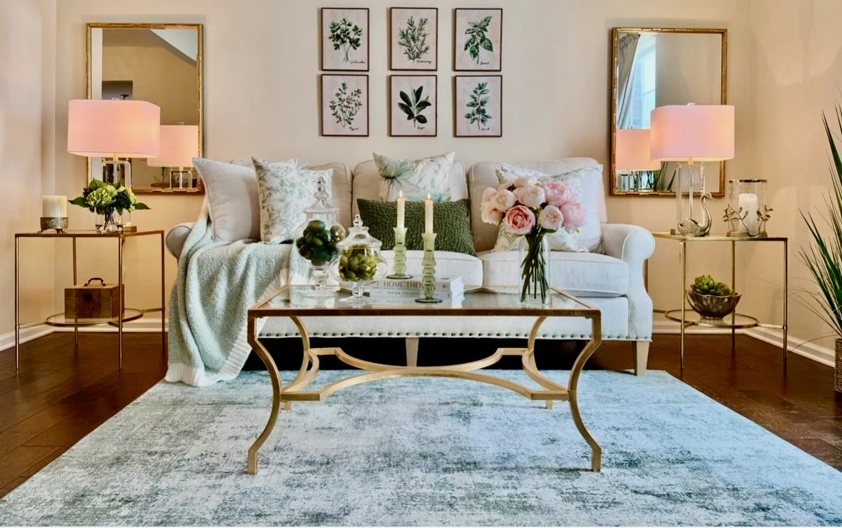 Stunning green and white living room styled with patterned sage green pillows, white solid pillows, a collection of 6 green framed prints over the couch. Two gold mirrors above the gold glass side tables styled with glass and gold lamps, green floral