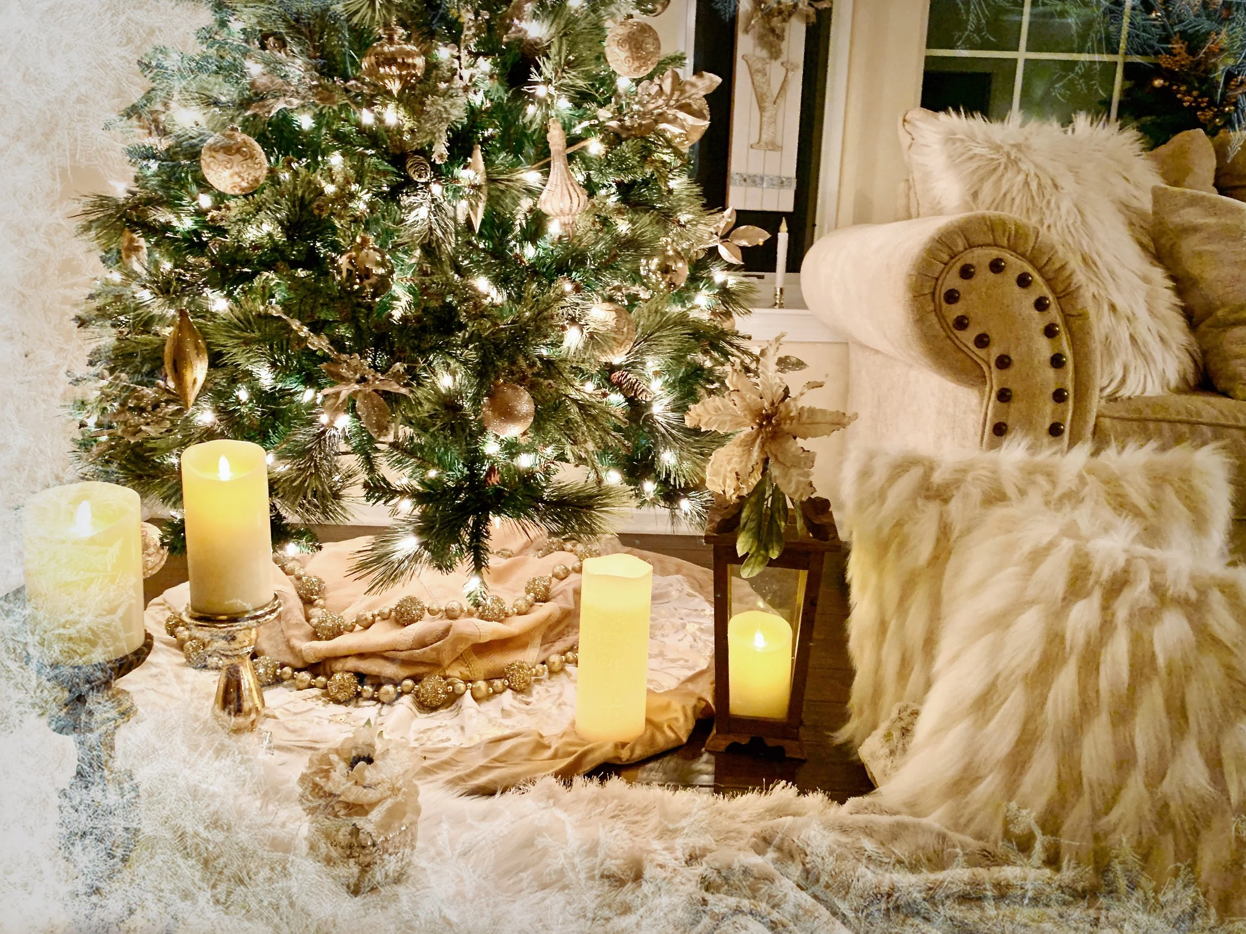 A decorated Christmas tree with gold ornaments and lights, surrounded by candles, a furry blanket, a pillow, and a floral vase in a cozy living room.