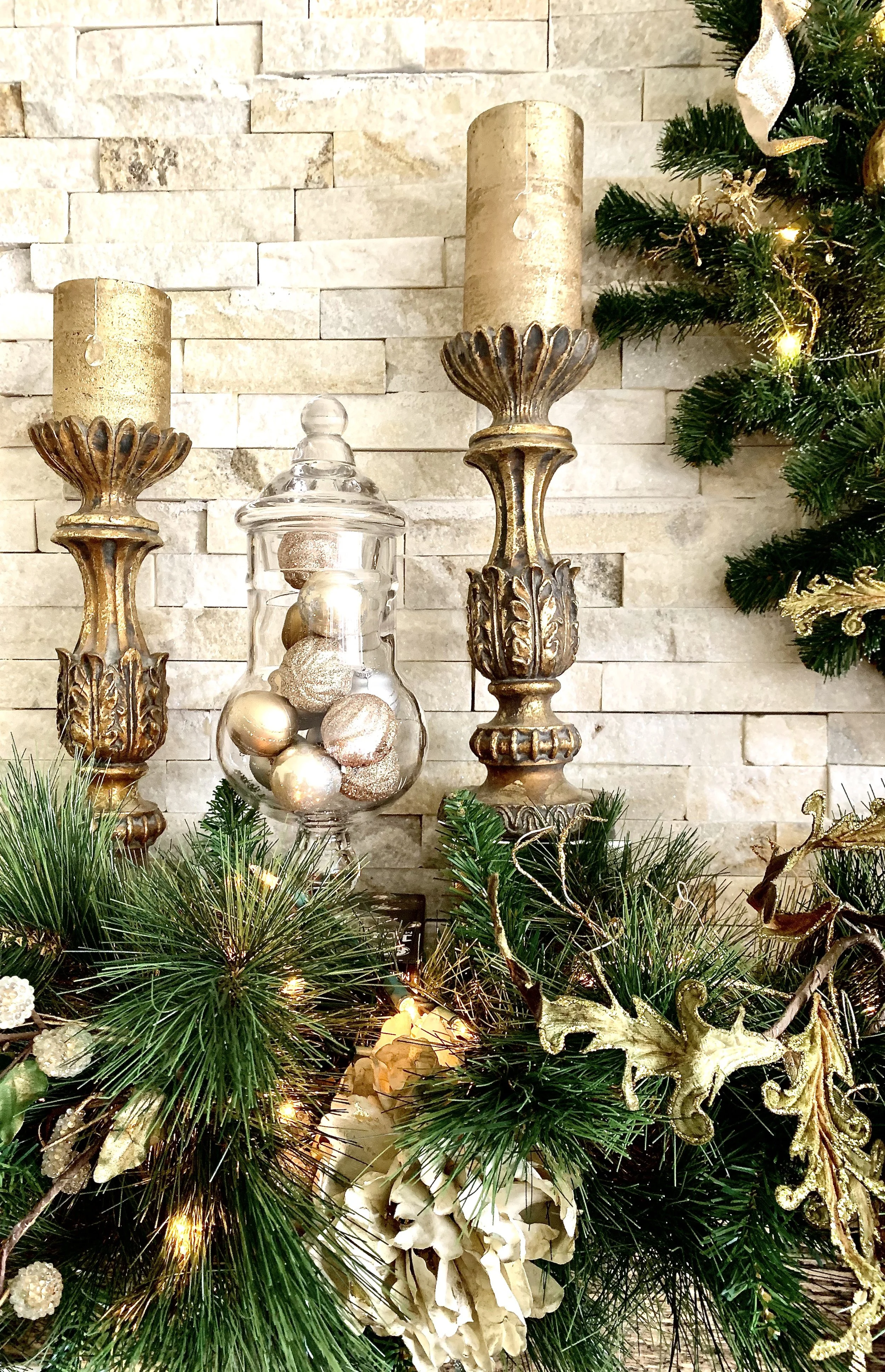 Decorative Christmas setup featuring gold pillar candles on ornate candle holders, a glass jar filled with gold and beige ornaments, and green pine branches with gold accents and lights, against a beige brick wall.