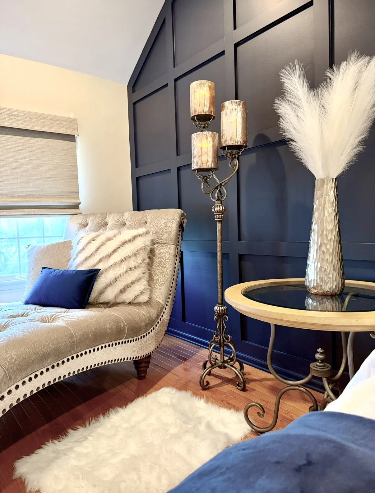 Living room corner with beige tufted chaise lounge, navy pillow, white fur pillow, and a window with beige shade. Decor includes a tall candle-style floor lamp with four lights, a cream textured vase with white pampas grass, a small glass-topped side
