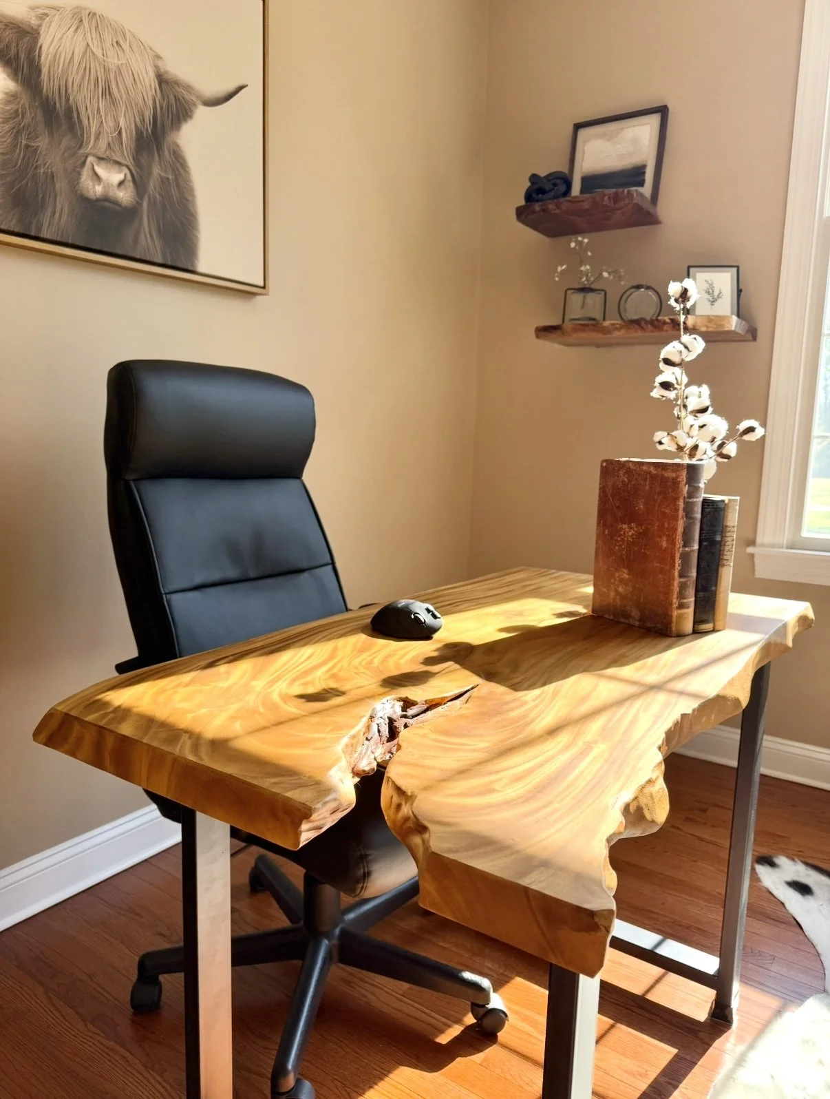 A home office with a live edge wooden desk, a black office chair, a desk mouse, a stack of vintage books, a jar with cotton stems, and wall-mounted shelves with framed art and decor, lit by sunlight from a nearby window.