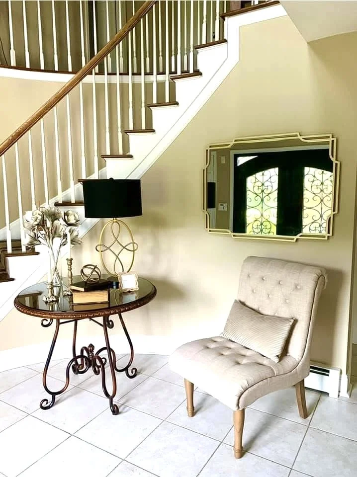 A cozy corner in a home featuring a beige tufted armchair with a matching pillow, a black and gold side table with decorative items, a classic, gold-framed mirror, a vase of flowers, and a black table lamp. The scene is near a staircase with white ba