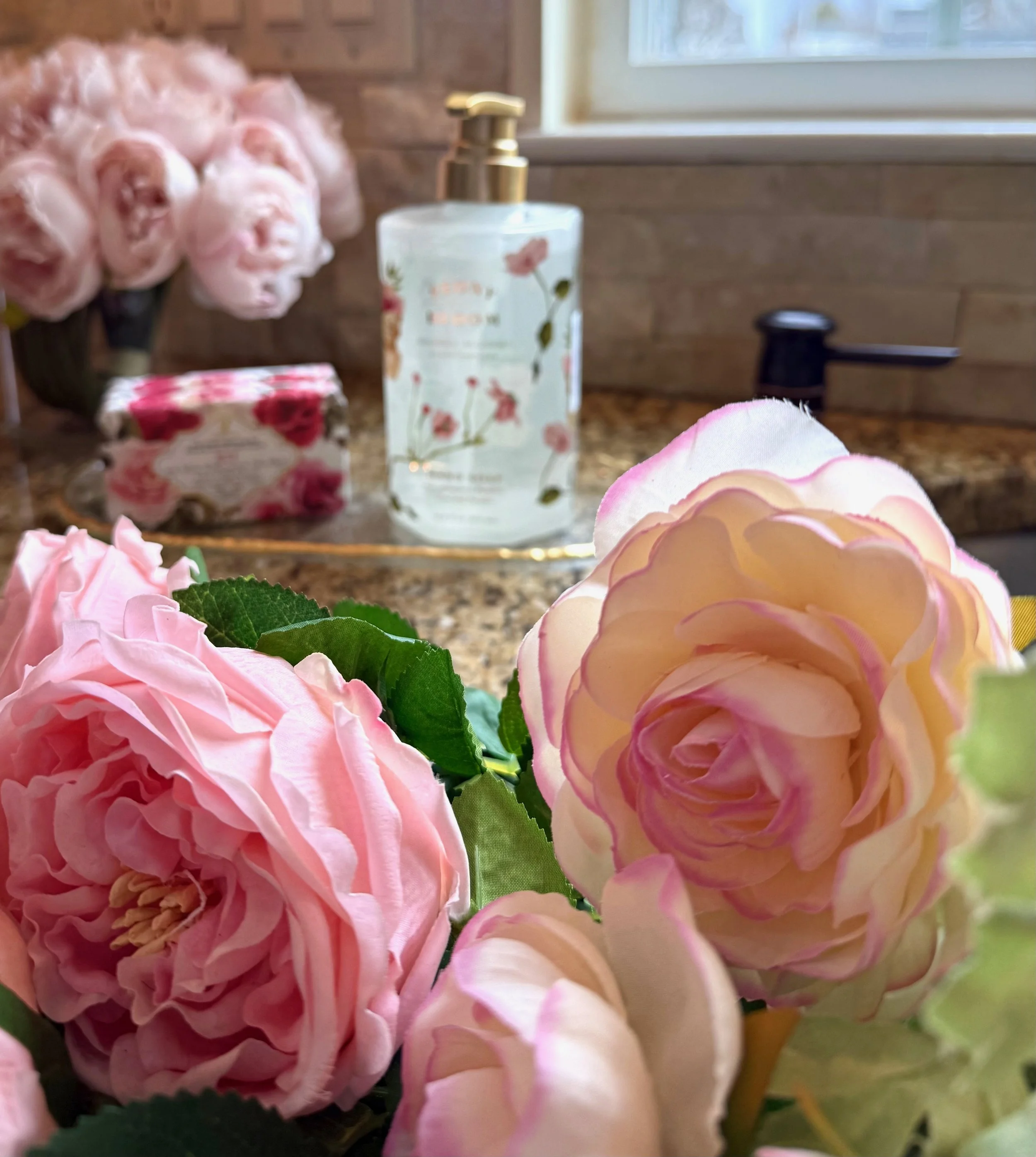 styled kitchen counter top and sink with pink flowers, a gold and glass tray holding a glass bottle hand soap and bars of rose decorated soap